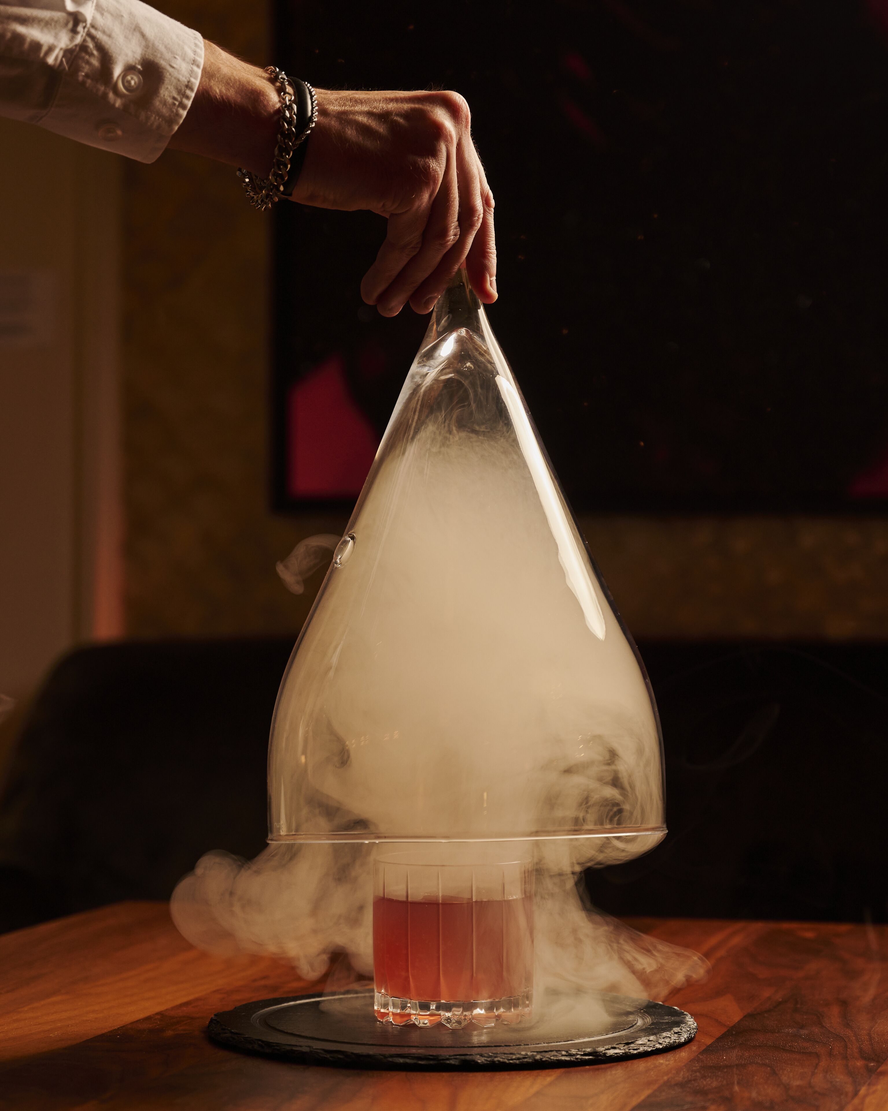 A hand holding a transparent glass dome over a drink on a coaster.