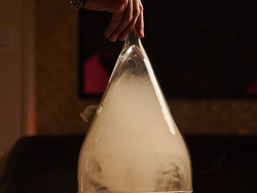 A hand holding a transparent glass dome over a drink on a coaster.