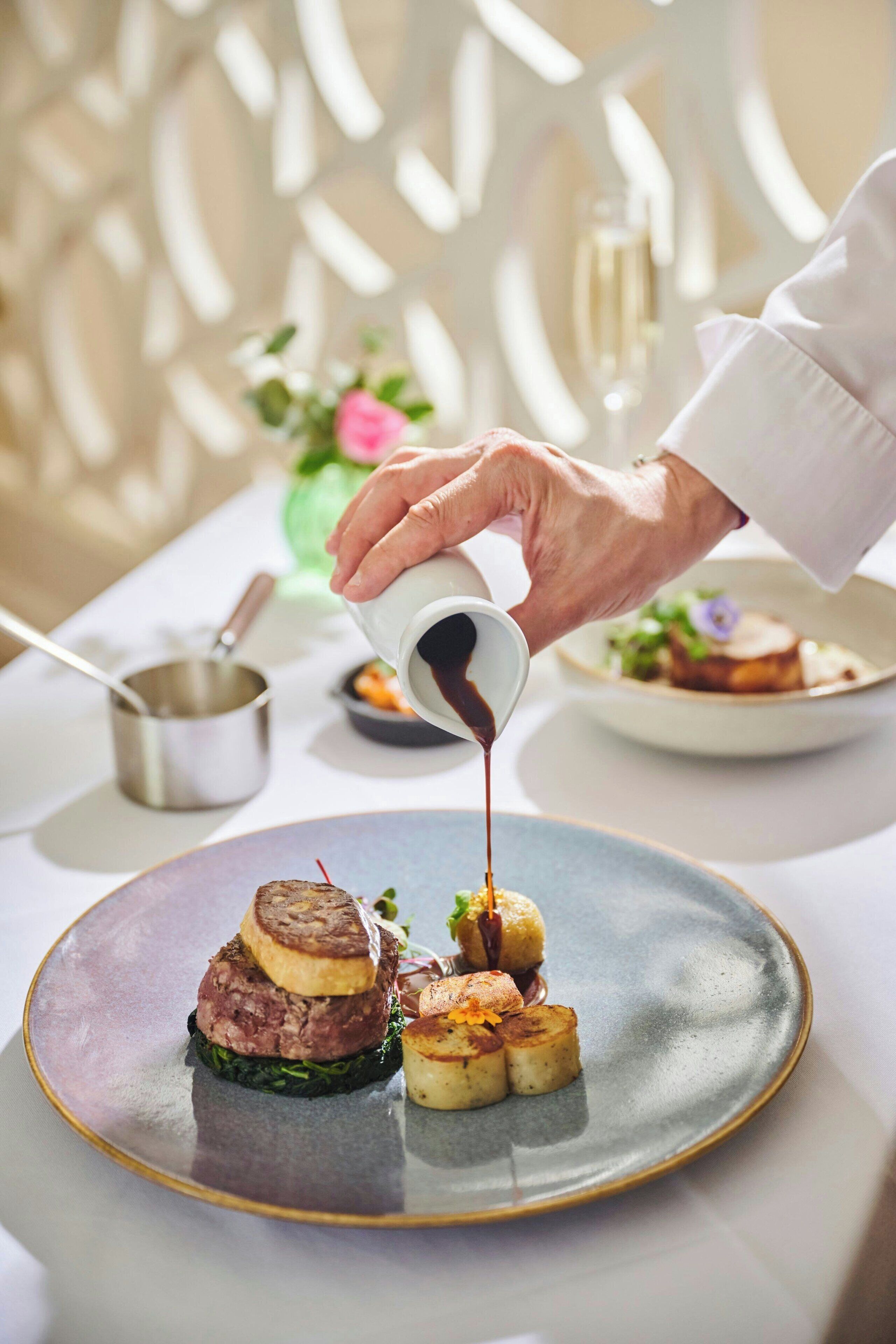 A person pouring sauce over a plated dish with steak and scallops.