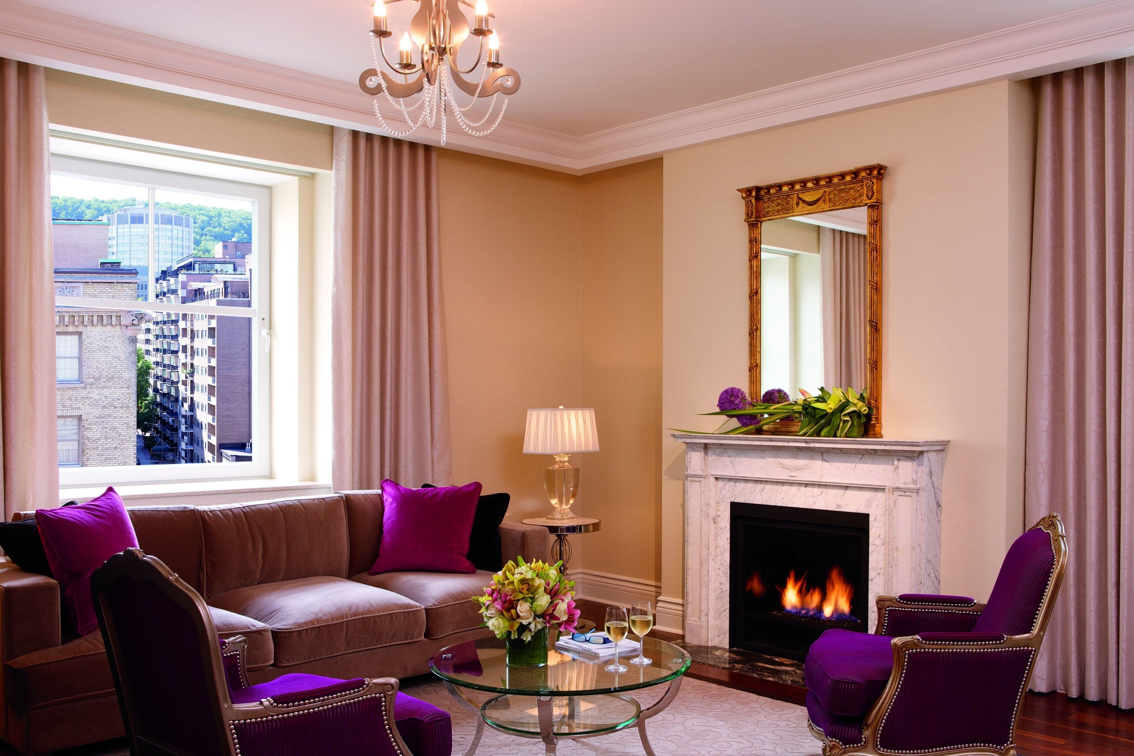 A living room with a sofa, armchairs, a glass coffee table, a fireplace, and a large window with curtains.