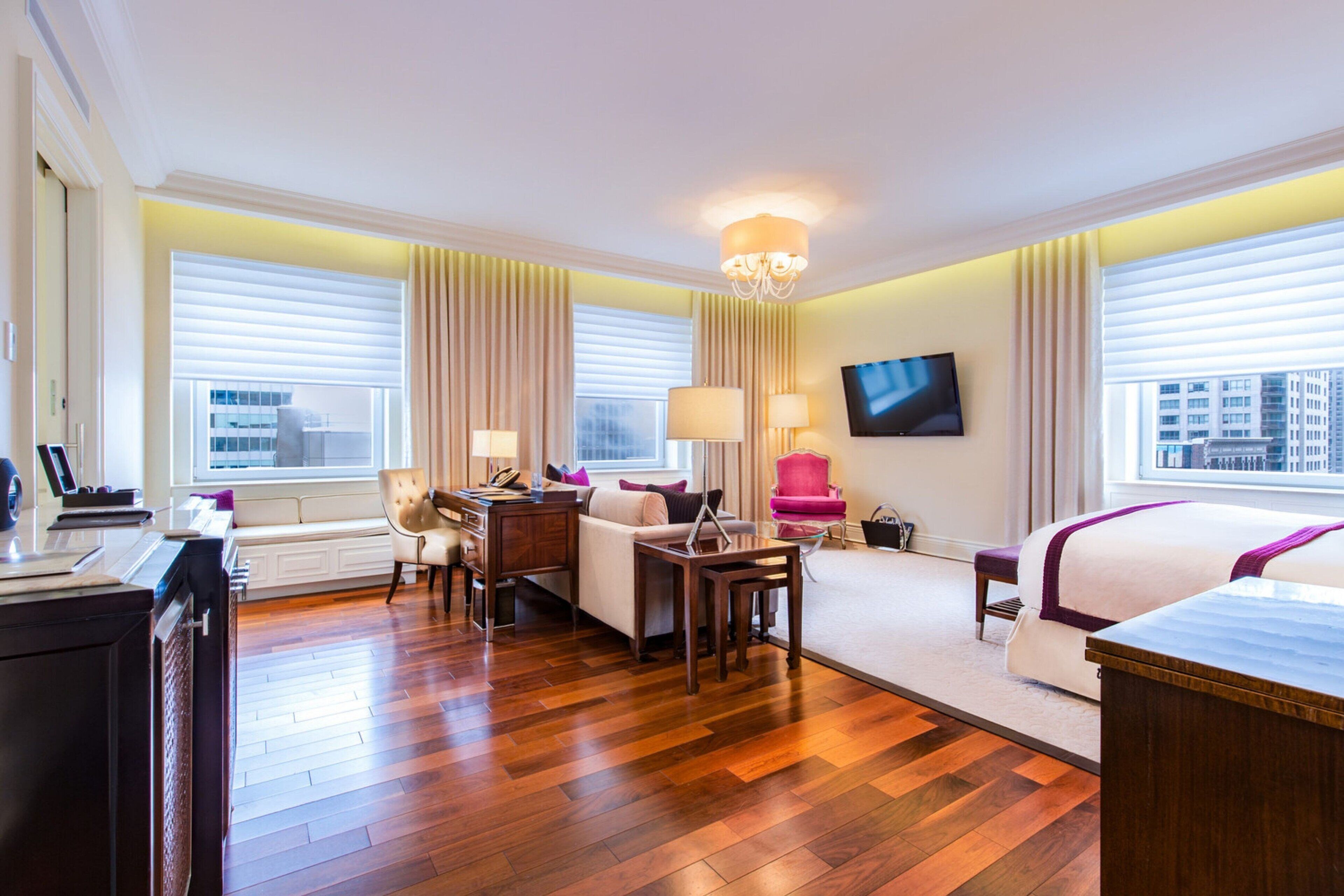 Habitación de hotel con piso de madera, un escritorio con una computadora portátil, un sofá, una silla, un televisor montado en la pared y ventanas amplias con cortinas.