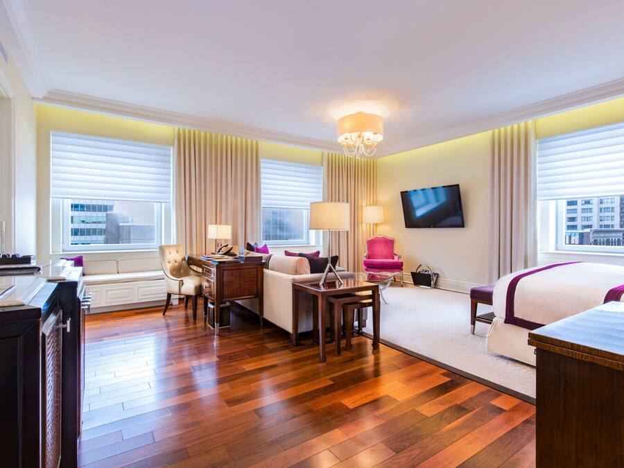 Ein Hotelzimmer mit Holzboden, einem Schreibtisch mit Laptop, einer Couch, einem Sessel, einem an der Wand befestigten Fernseher und großen Fenstern mit Vorhängen.
