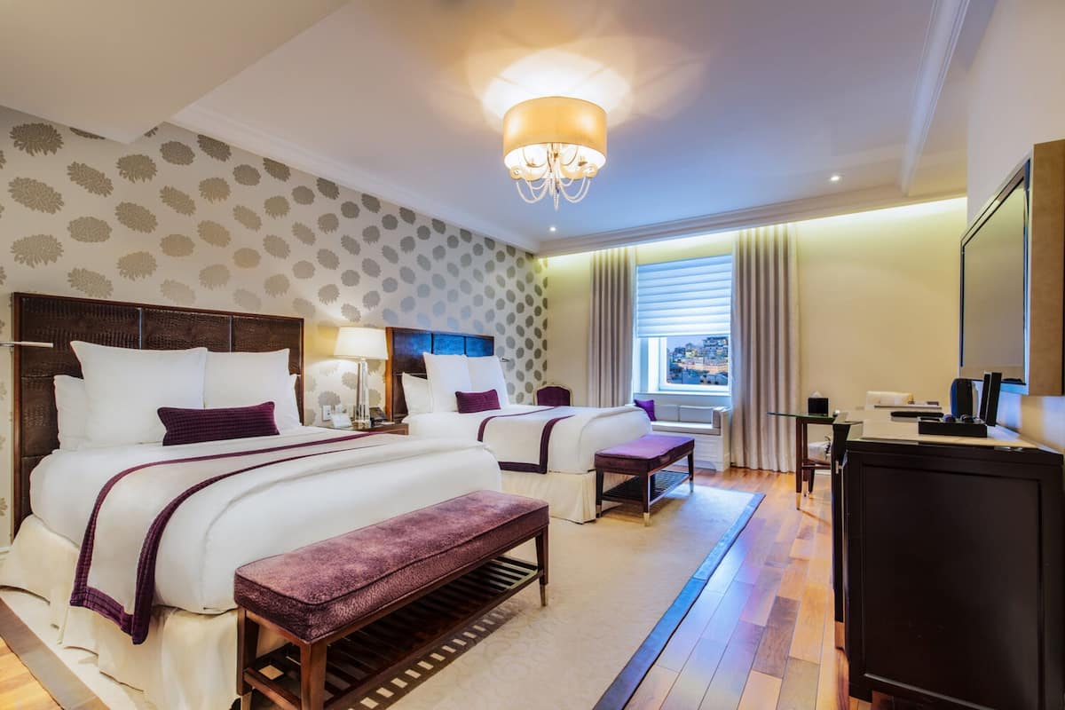 A hotel room with two beds, a desk, a chair, a bench, a TV, and a window with curtains.