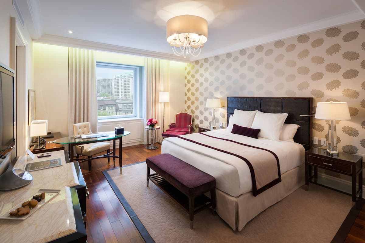 A hotel room with a large bed, bedside tables, a desk with a chair, a TV, and a window with a view of buildings.