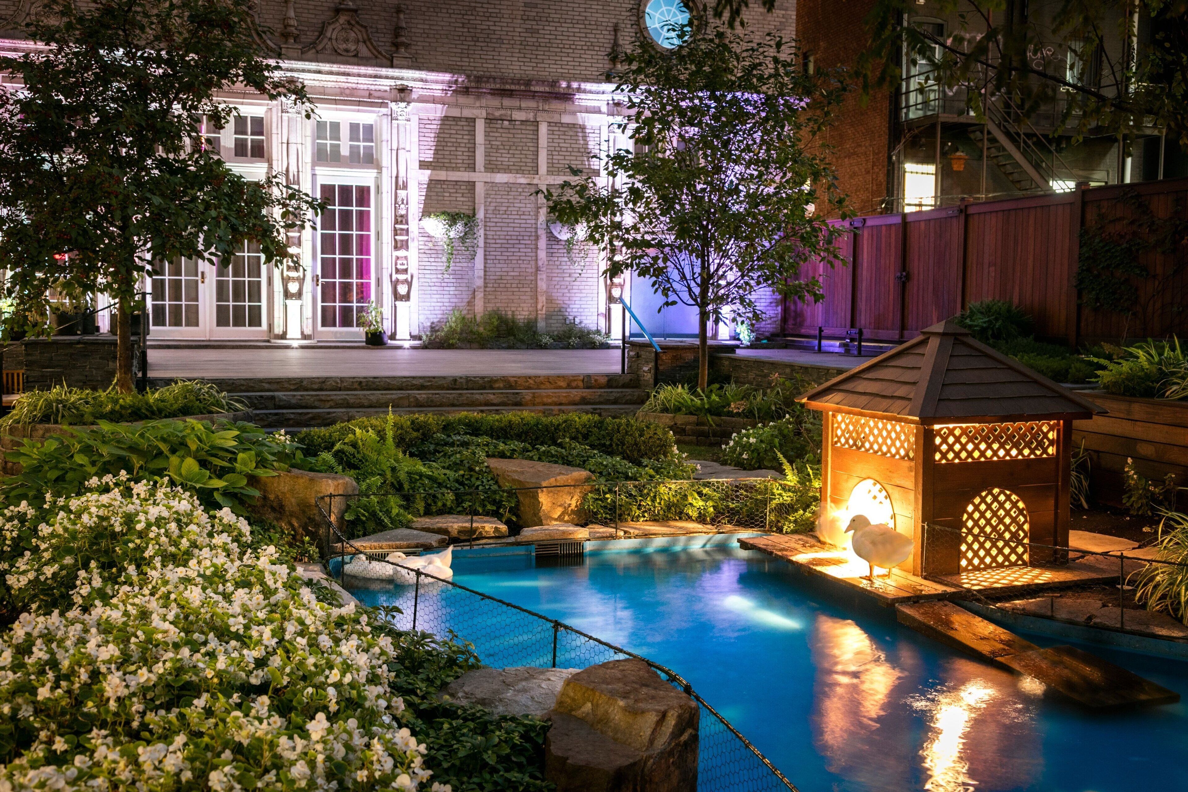 A nighttime pool area with a wooden deck, a small gazebo, and a brick building in the background.