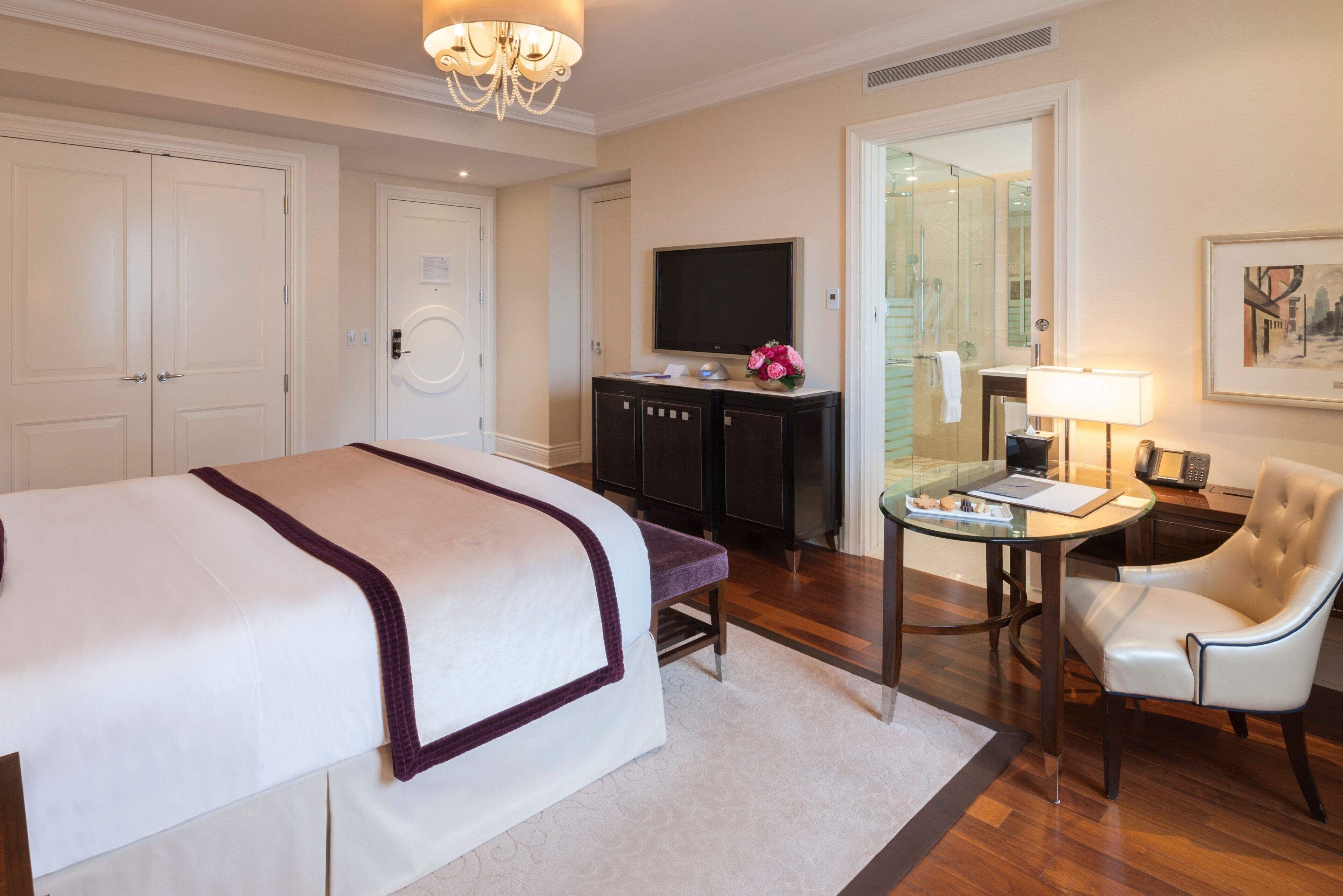 A hotel room with a large bed, a desk with a chair, a television, and a bathroom visible through a glass door.