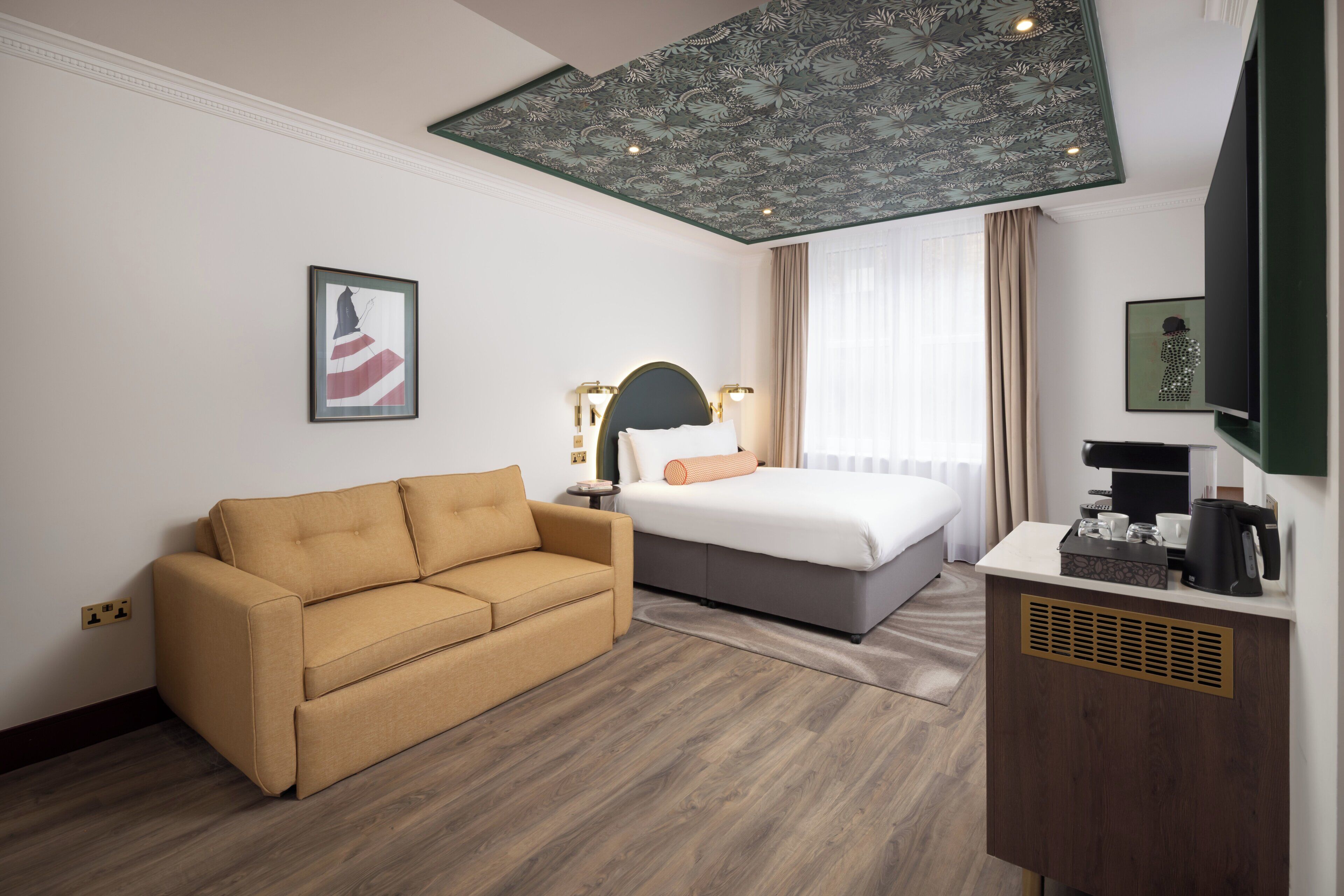 A hotel room with a bed, a sofa, a desk with a coffee machine, and a ceiling with a floral pattern.
