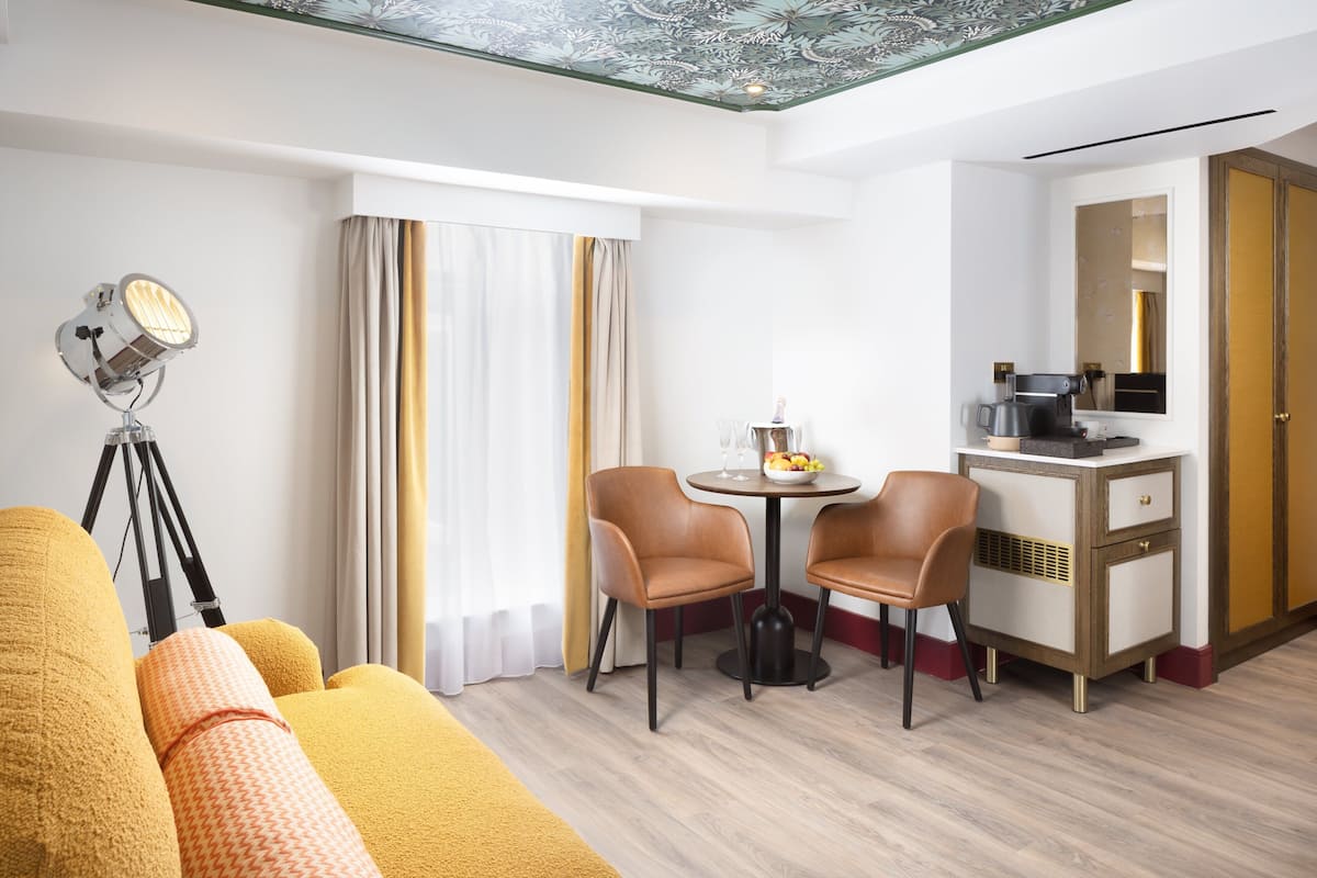 A hotel room with a sofa, two chairs, a small table with a fruit bowl, a desk with a computer, and a tripod.