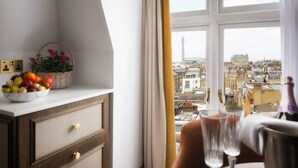 Ein Zimmer mit Blick auf eine Stadtlandschaft durch ein Fenster, ein Esstisch mit einer Champagnerflasche und eine Obstschale auf einer Kommode.