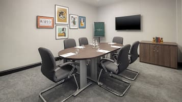 A conference room with a round table, six chairs, a flat-screen TV, framed artwork, and a wooden cabinet.