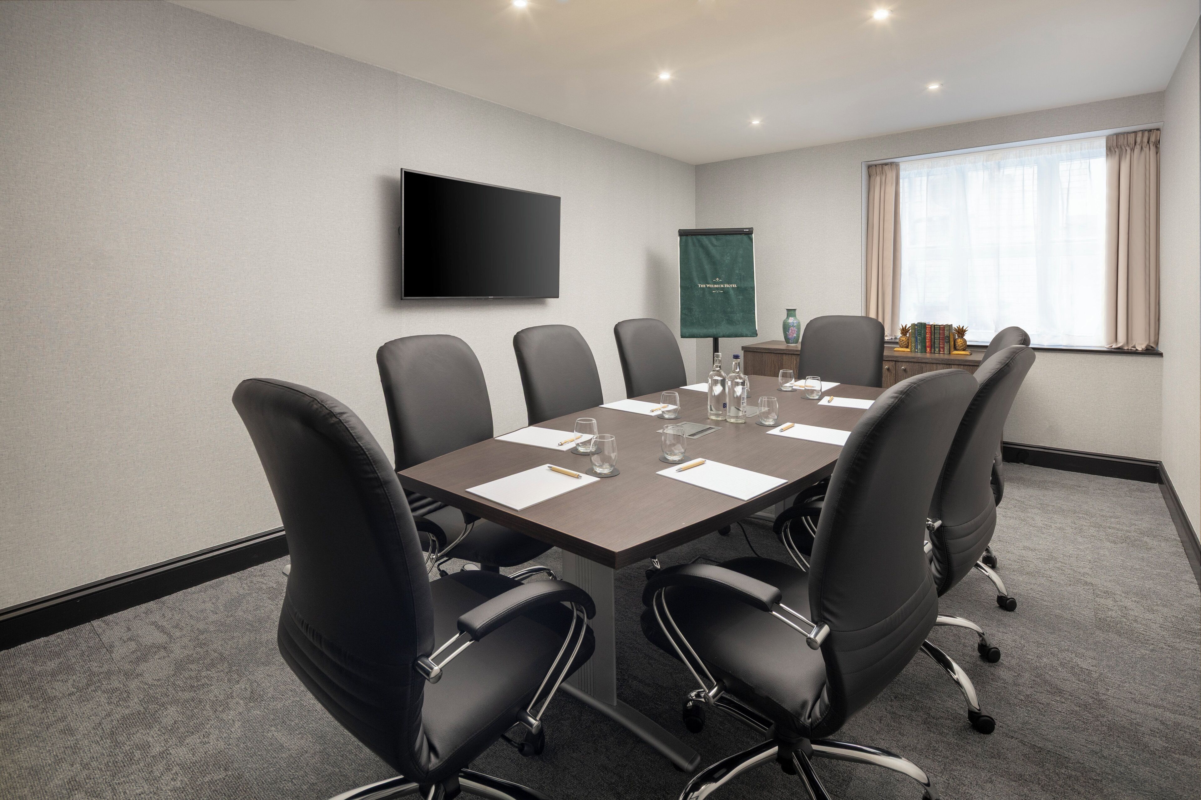 A conference room with a large table, six chairs, a flat-screen TV, a projector screen, and a window with curtains.