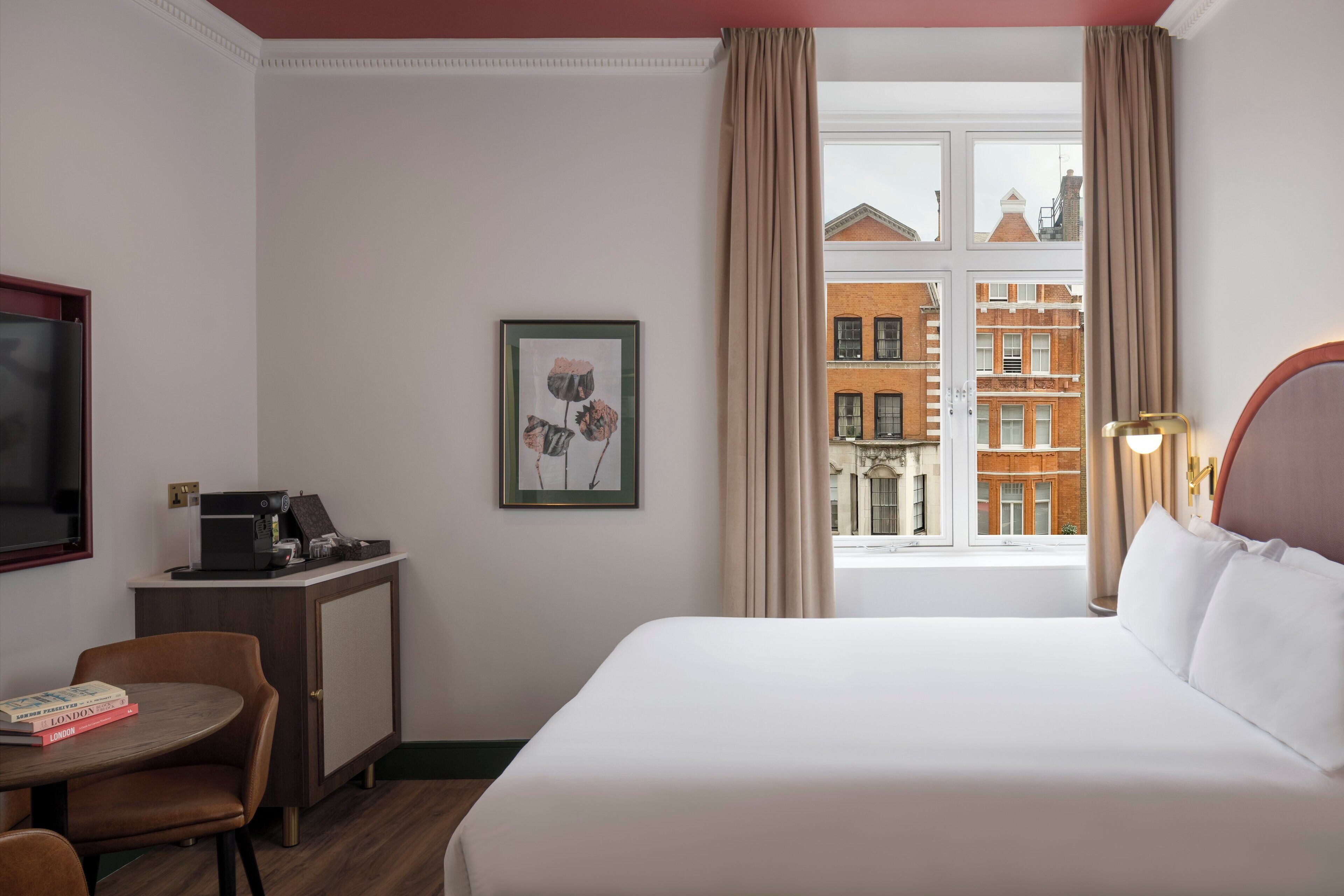 Habitación de hotel con cama, escritorio con cafetera, silla, cuadro enmarco y ventana con vista a edificios.