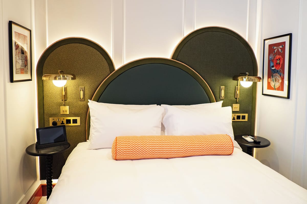 A hotel room with a large bed, two wall-mounted lamps, a laptop on a side table, and framed artwork on the walls.