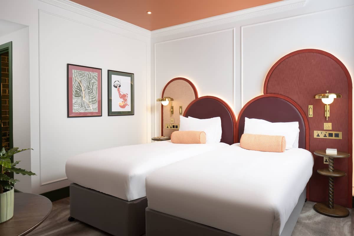 A hotel room with two beds, a potted plant, framed artwork on the wall, and a round table with a lamp.