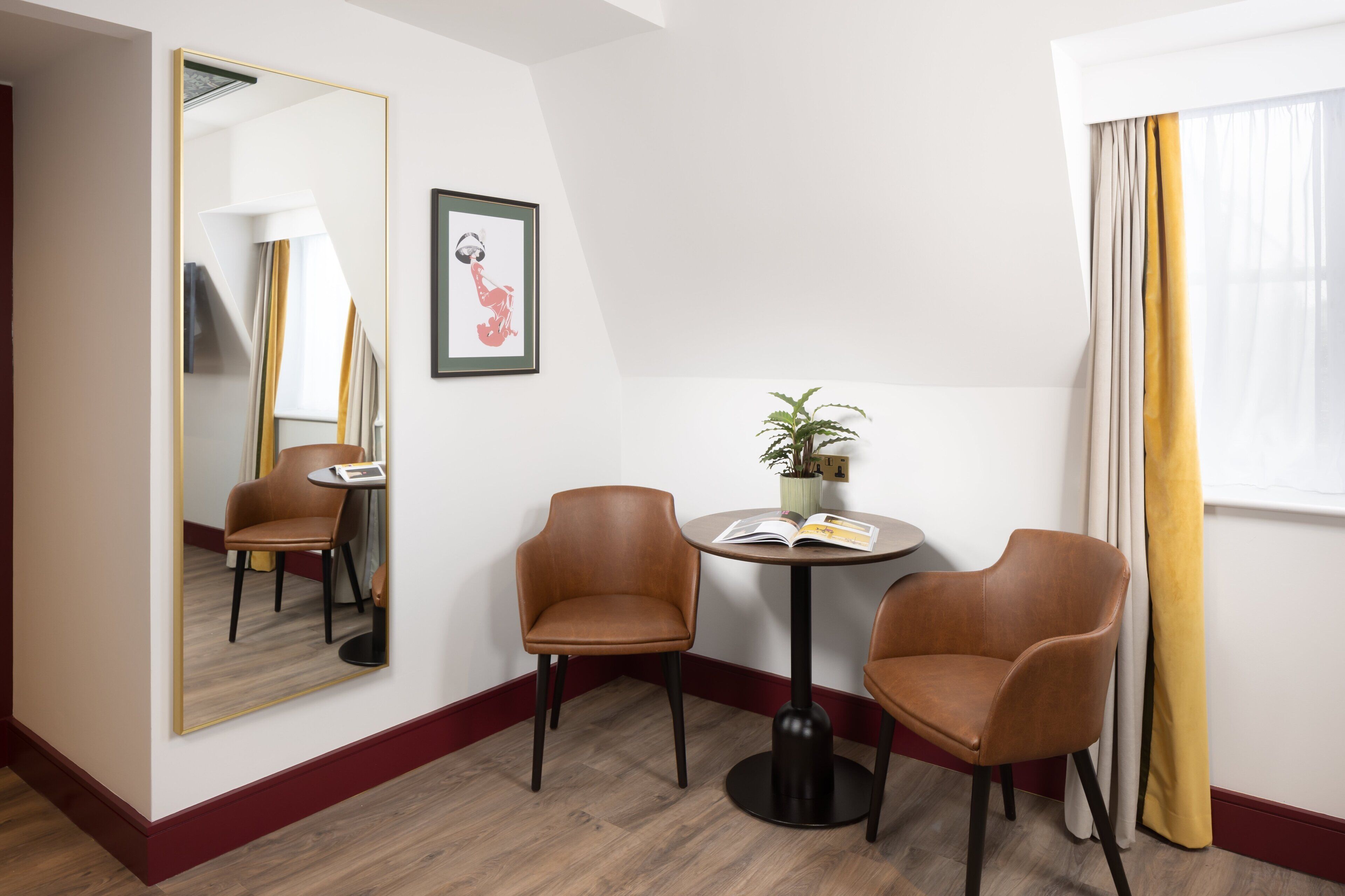 A hotel room with a large mirror, two brown leather chairs, a round table with a book and a plant, and a window with yellow curtains.