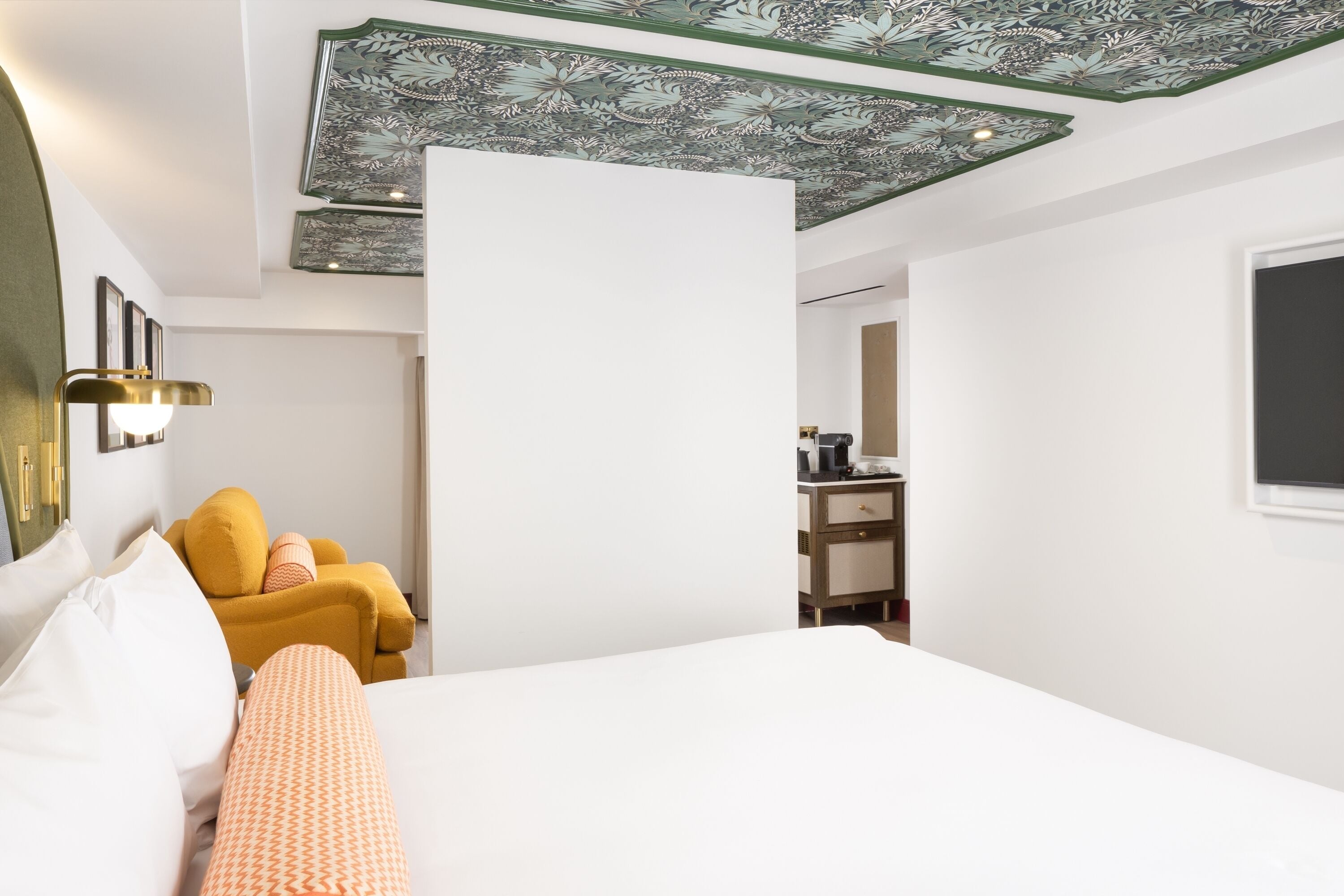 A hotel room with a bed, a yellow armchair, a desk with a chair, and a mirror on the wall.