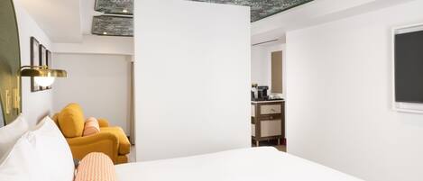 A hotel room with a bed, a yellow armchair, a desk with a chair, and a mirror on the wall.
