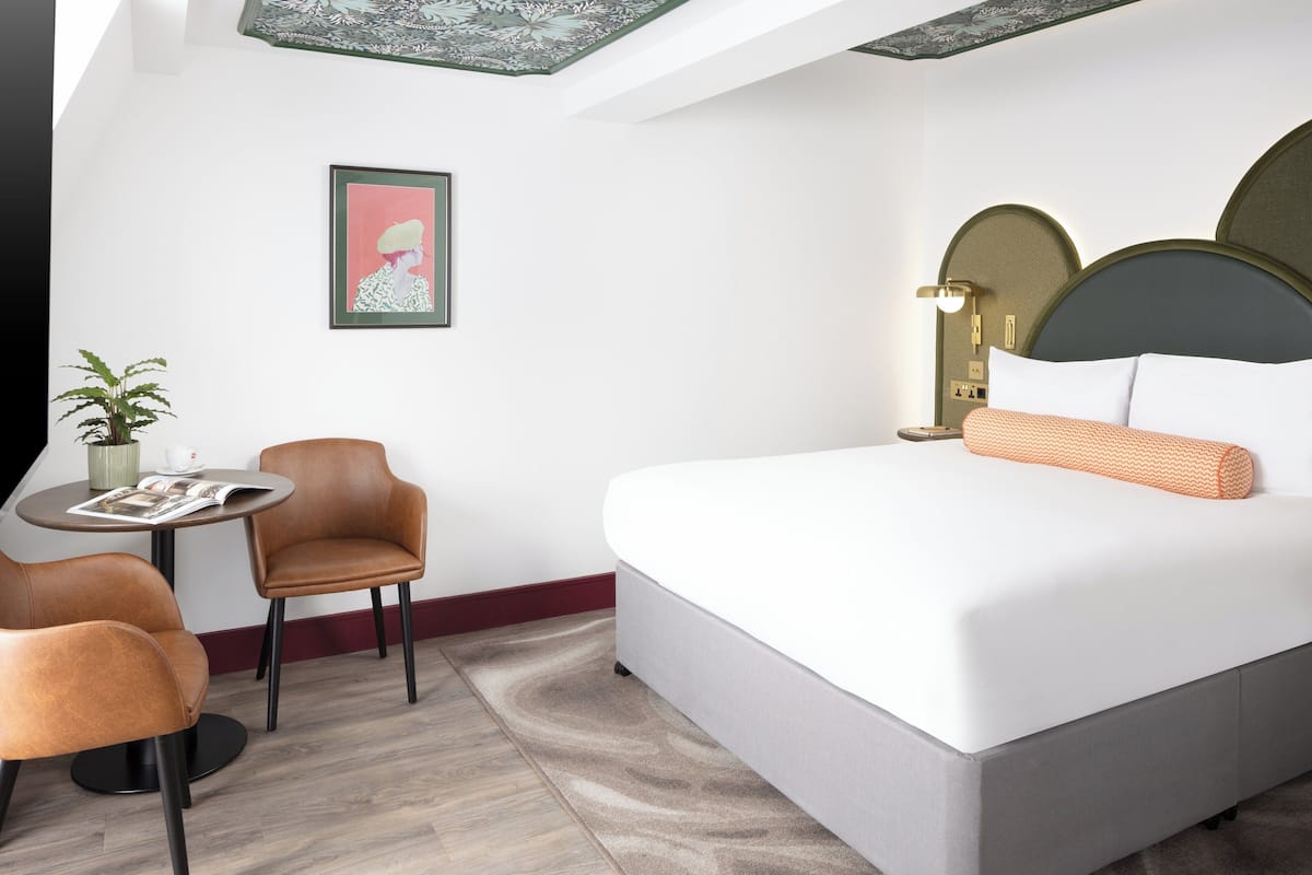 A modern hotel room with a large bed, a round table with two chairs, a potted plant, and a framed picture on the wall.