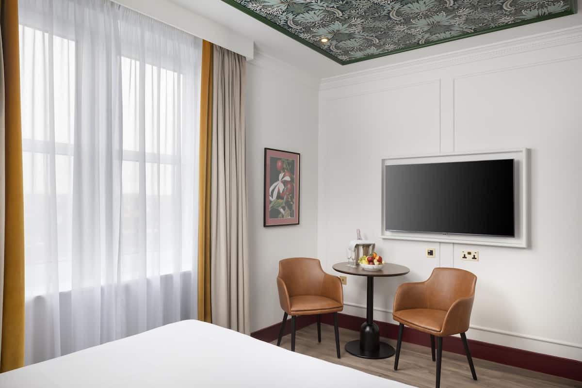 A hotel room with a bed, two chairs, a table with a fruit bowl, a flat-screen TV, and a painting on the wall.