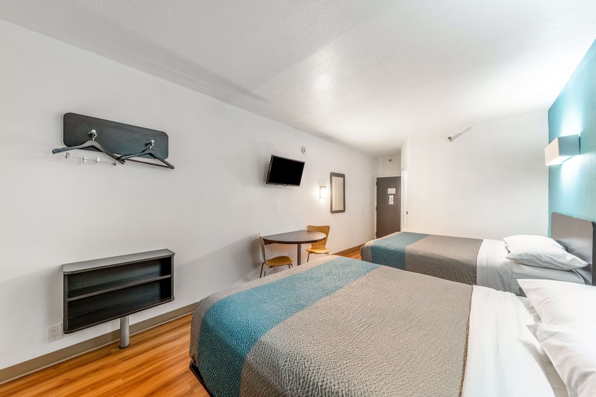 Ein Hotelzimmer mit zwei Betten, einem Fernseher, einem kleinen Tisch mit Stühlen und einem Kleiderhaken.