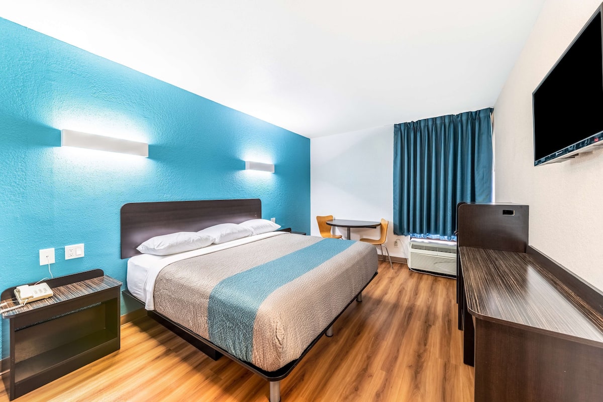 Ein Hotelzimmer mit Bett, Nachttisch, Schreibtisch, Stuhl und einem an der Wand befestigten Fernseher.