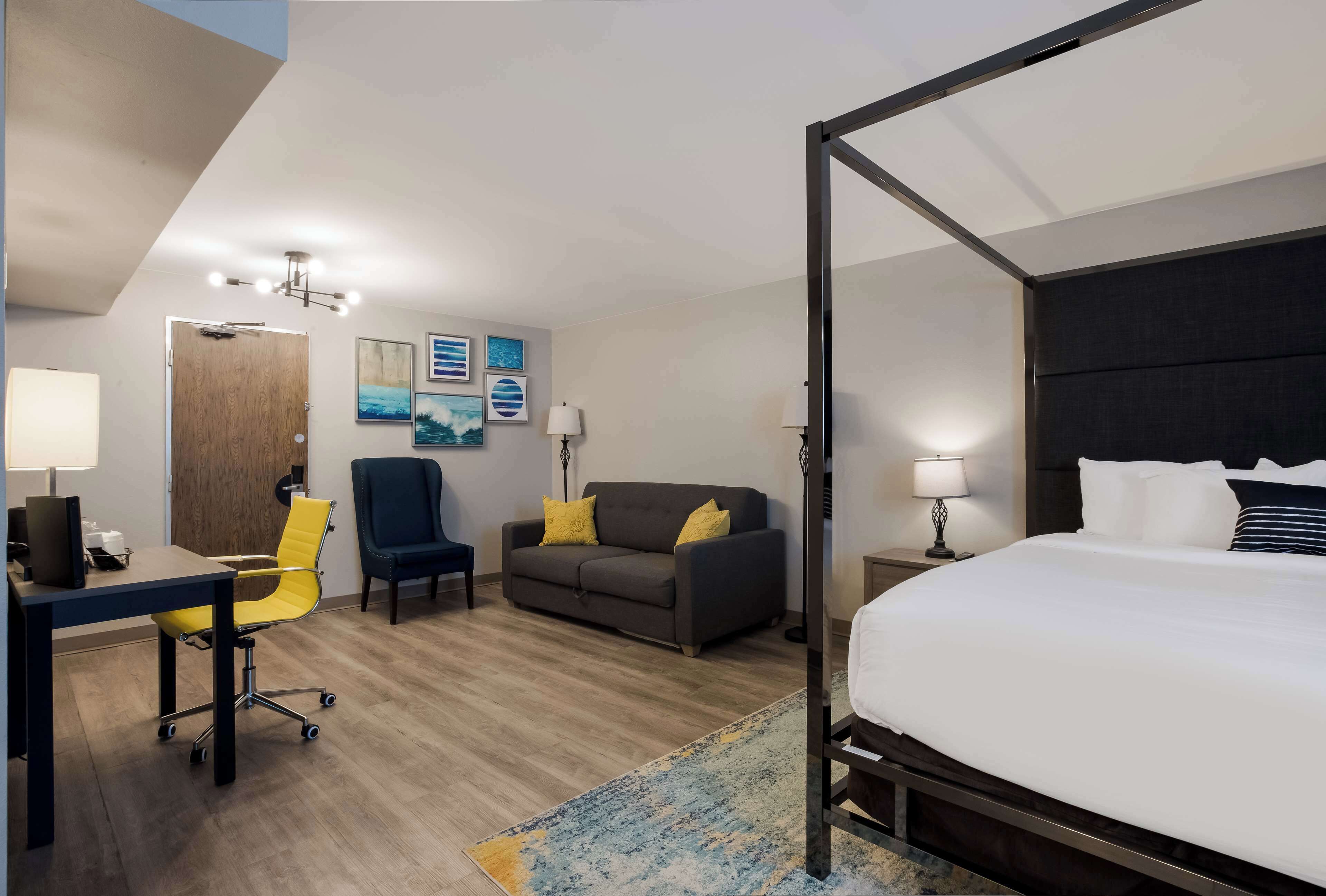 A modern hotel room with a four-poster bed, a desk with a chair, a sofa, and a small table with a lamp.