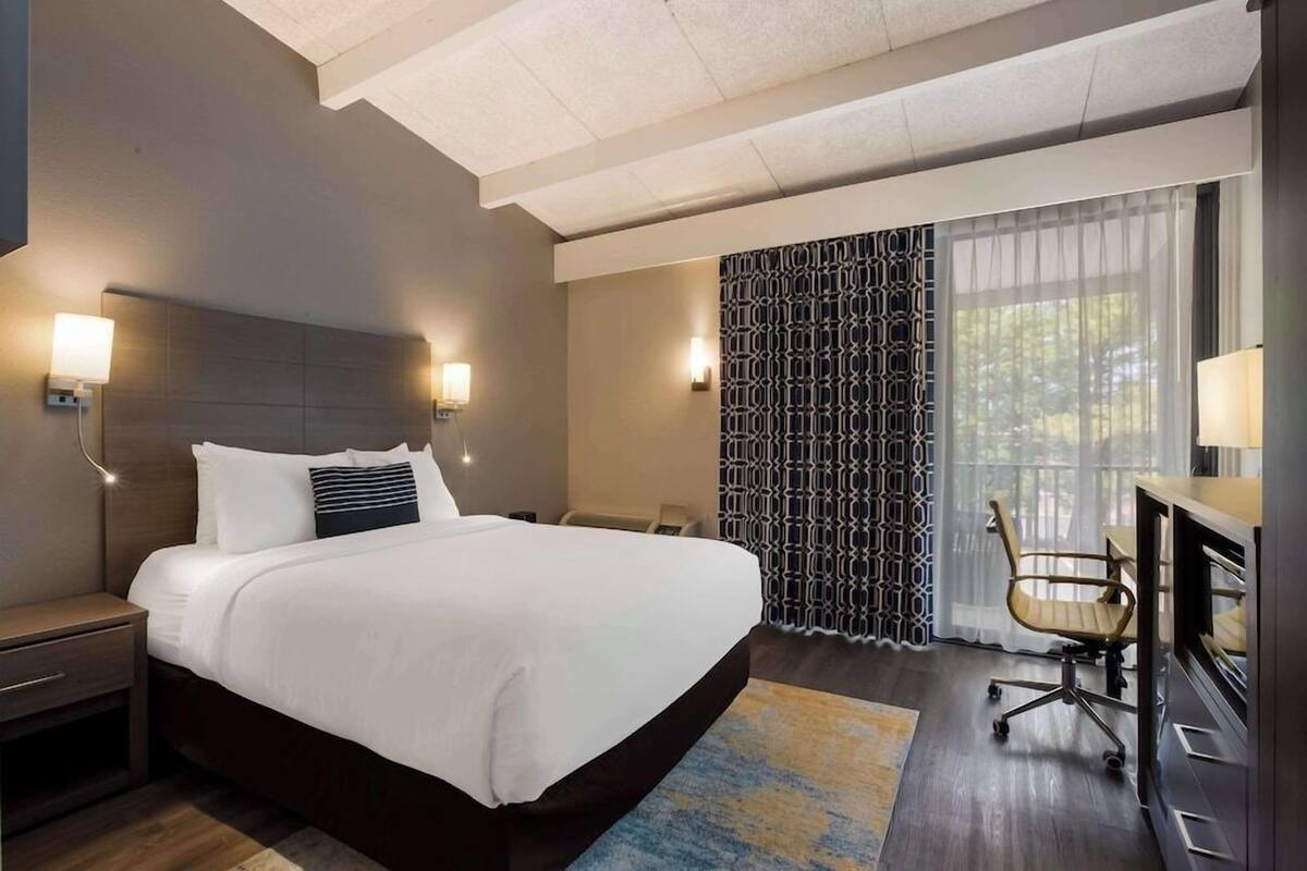 A hotel room with a large bed, bedside tables, a desk with a chair, a patterned curtain, and a view of the outdoors.