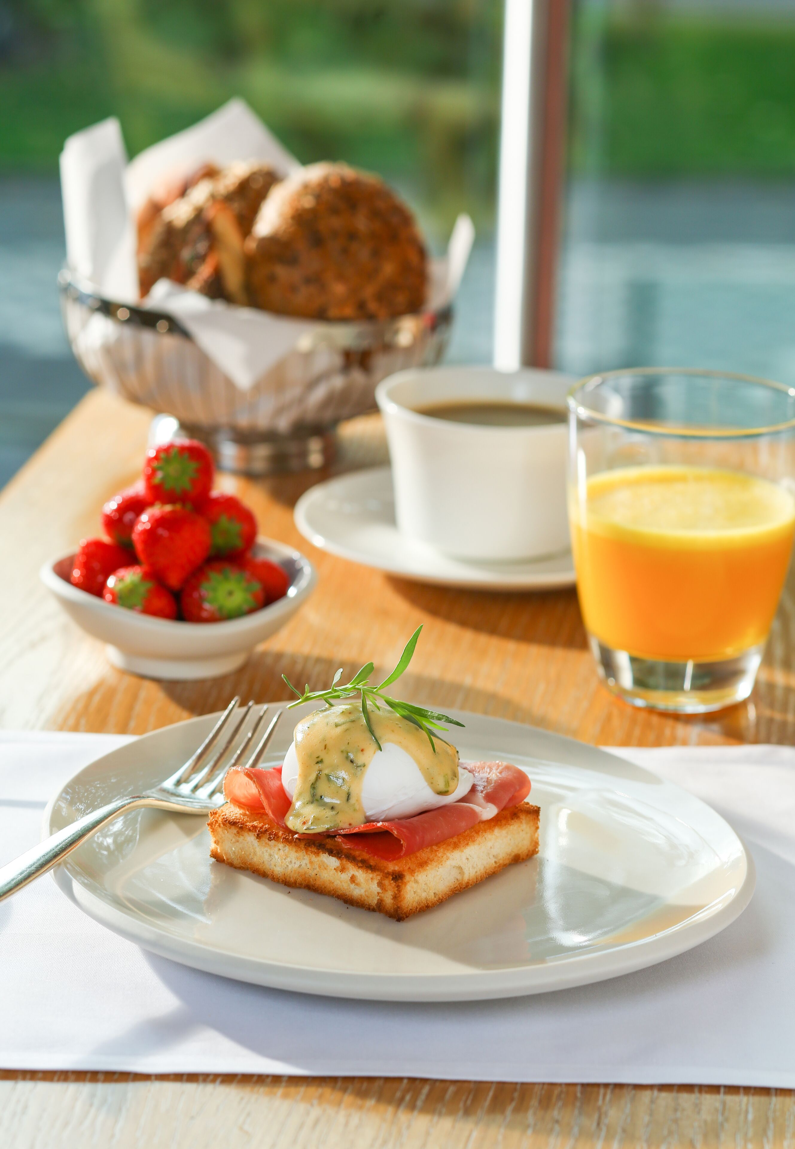 A breakfast setting with a slice of toast topped with smoked salmon and a poached egg, a bowl of strawberries, a glass of orange juice, and a cup of coffee on a wooden table.