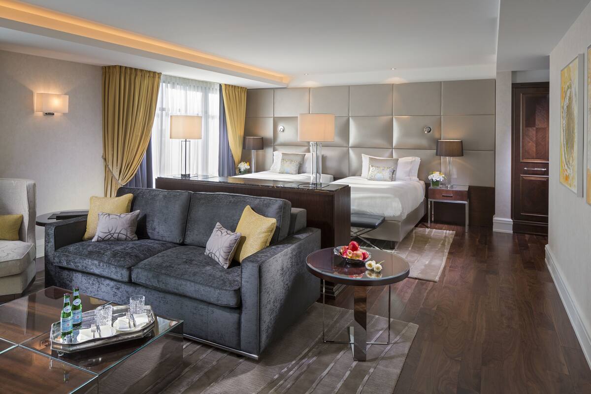 Una habitación de hotel moderna con un sofá gris, una cama con cabecera, una mesita de noche con lámparas y una mesa de centro de cristal con una bandeja de vasos y botellas.