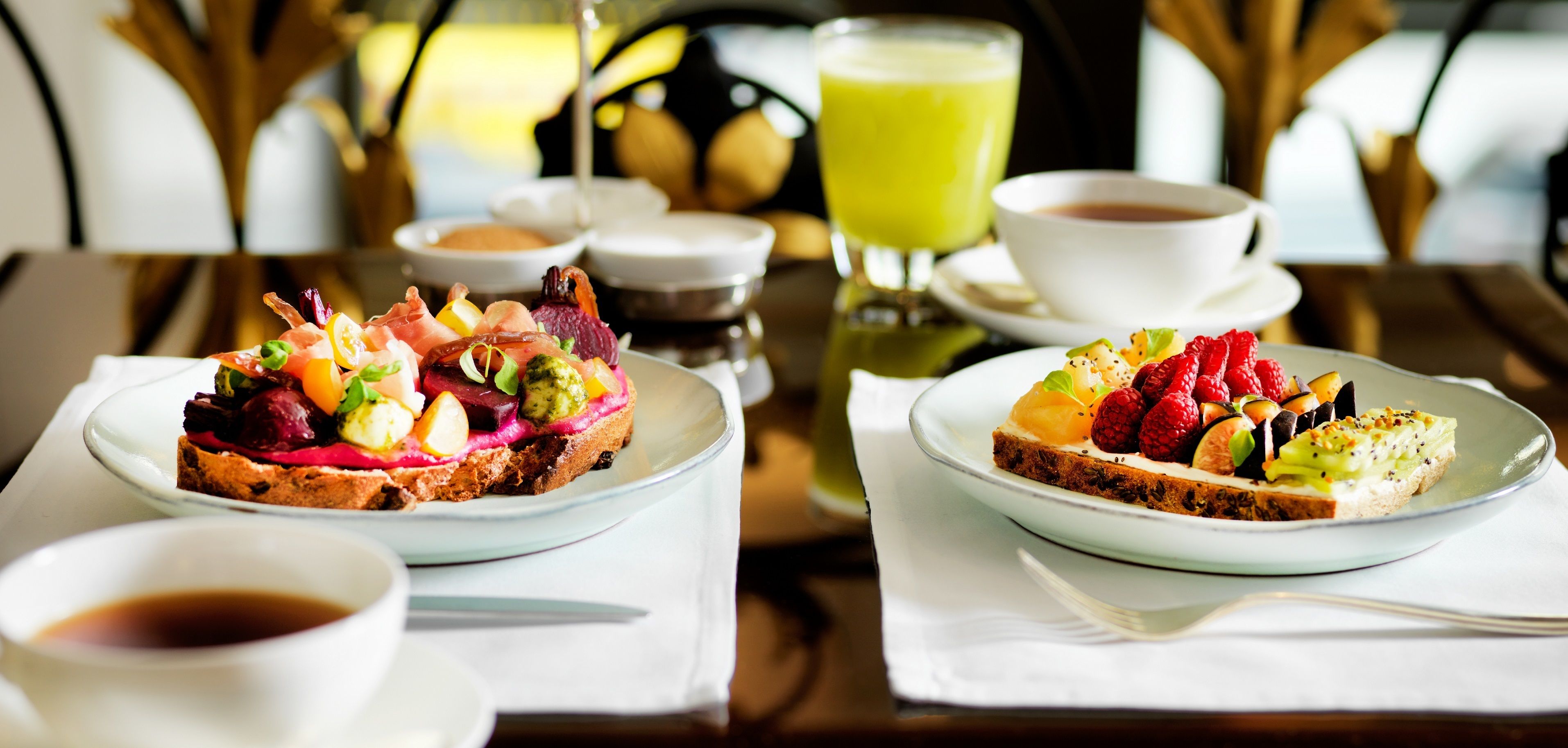 Dos tostadas con diferentes ingredientes, una taza de café, un vaso de jugo verde y un recipiente pequeño con salsa sobre una mesa.