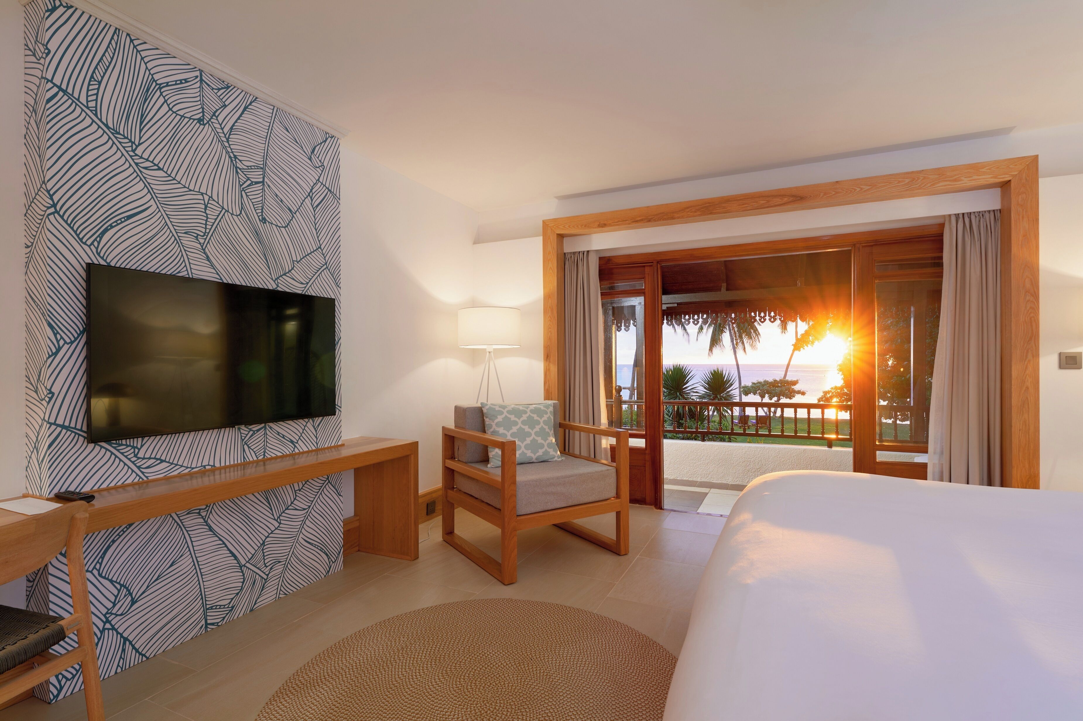 Habitación de hotel con televisión, escritorio, silla, cama y balcón con vista al atardecer.