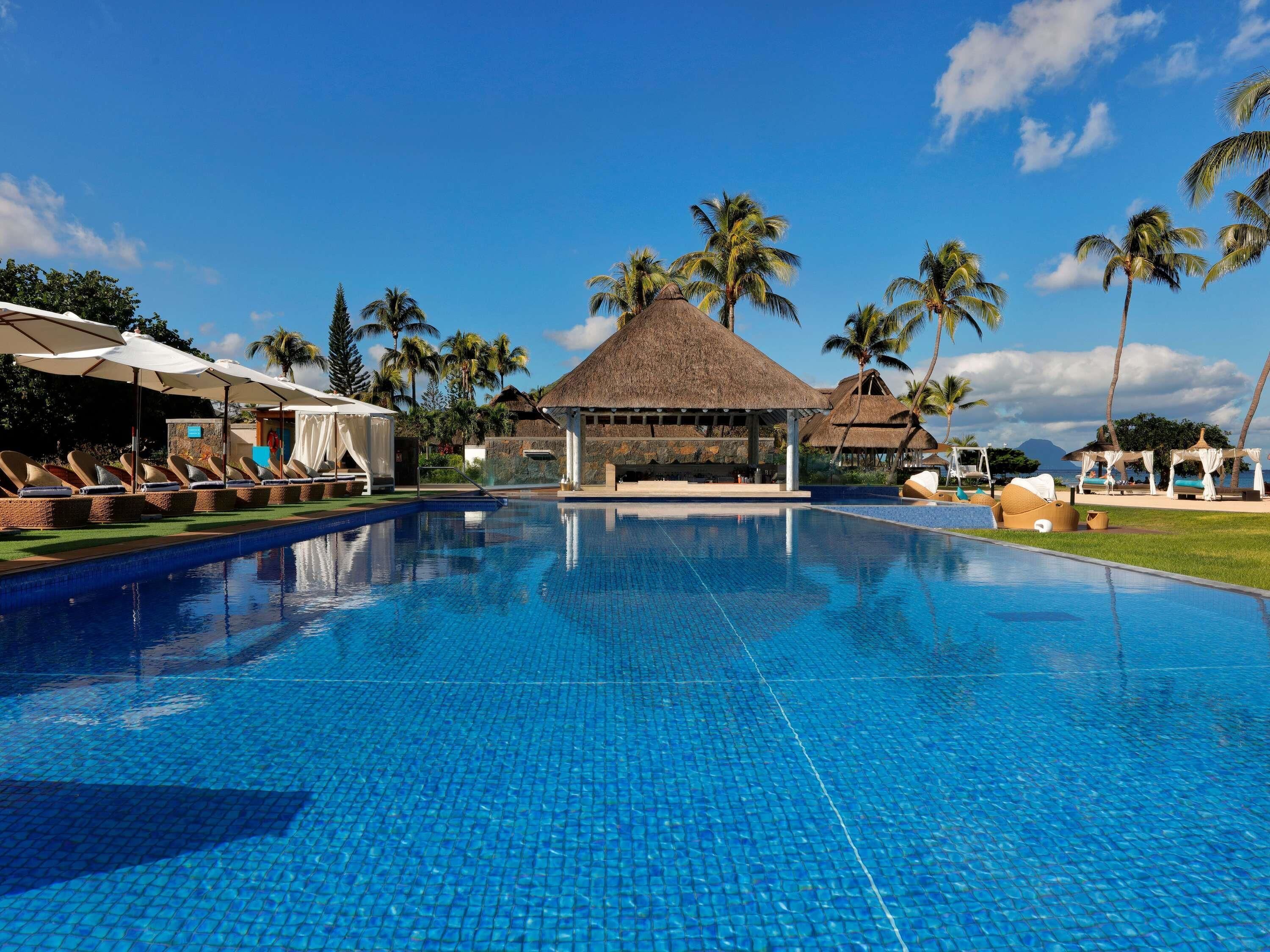 A swimming pool with lounge chairs and umbrellas, surrounded by palm trees and thatched-roof structures.