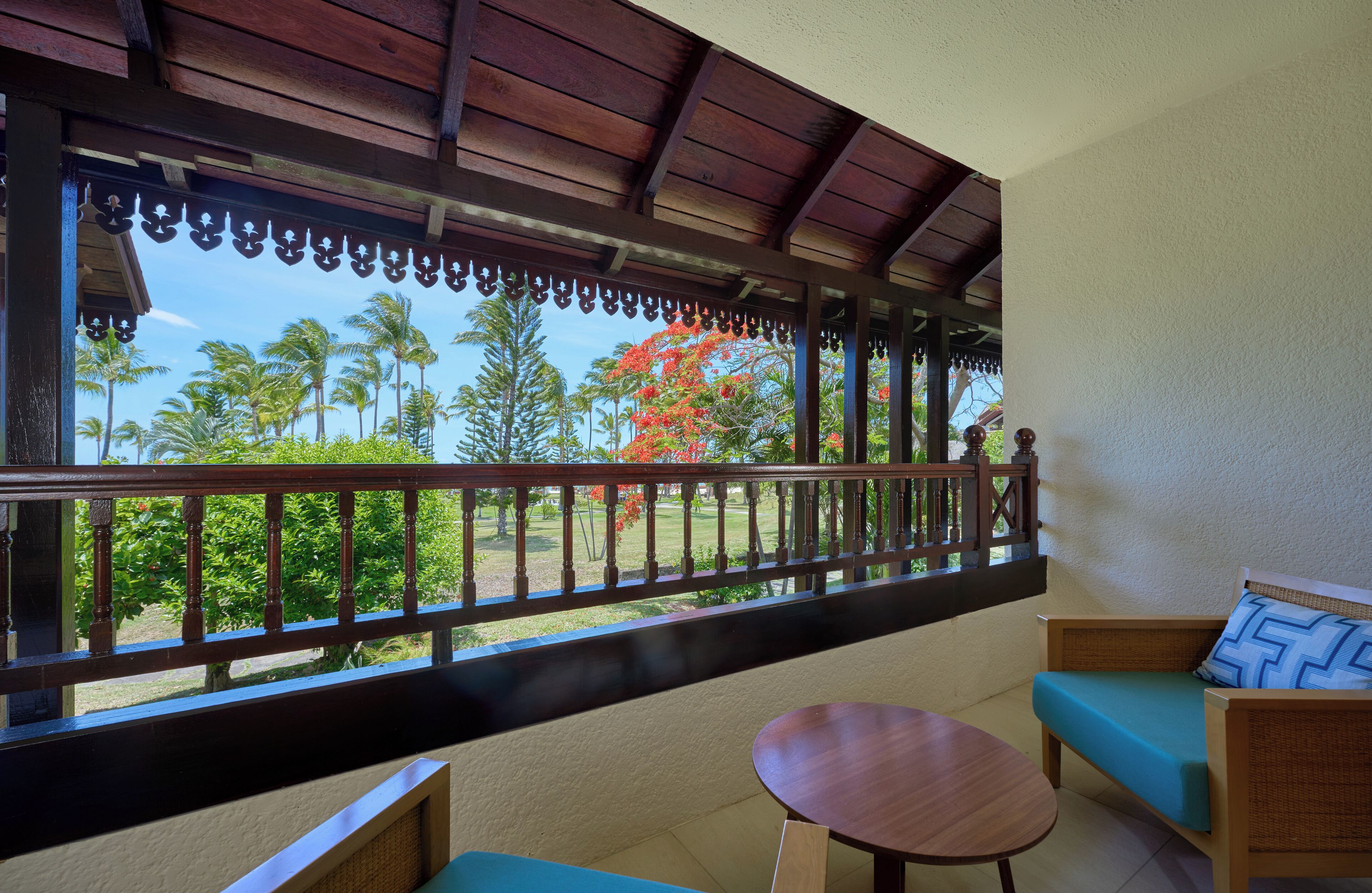 Un balcón con barandilla de madera, un sofá de mimbre, una mesa redonda de madera y vistas a palmeras y un árbol de flores rojas.