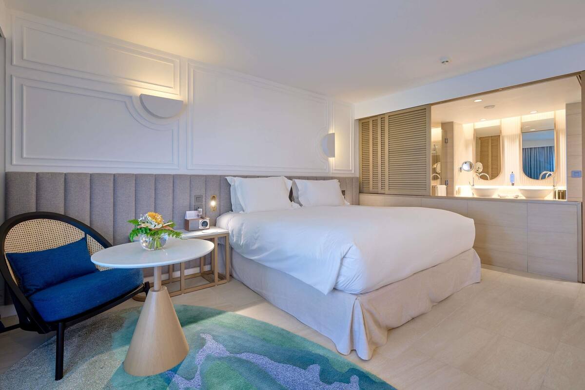 A hotel room with a bed, bedside table, chair, and a bathroom with a mirror and sink.