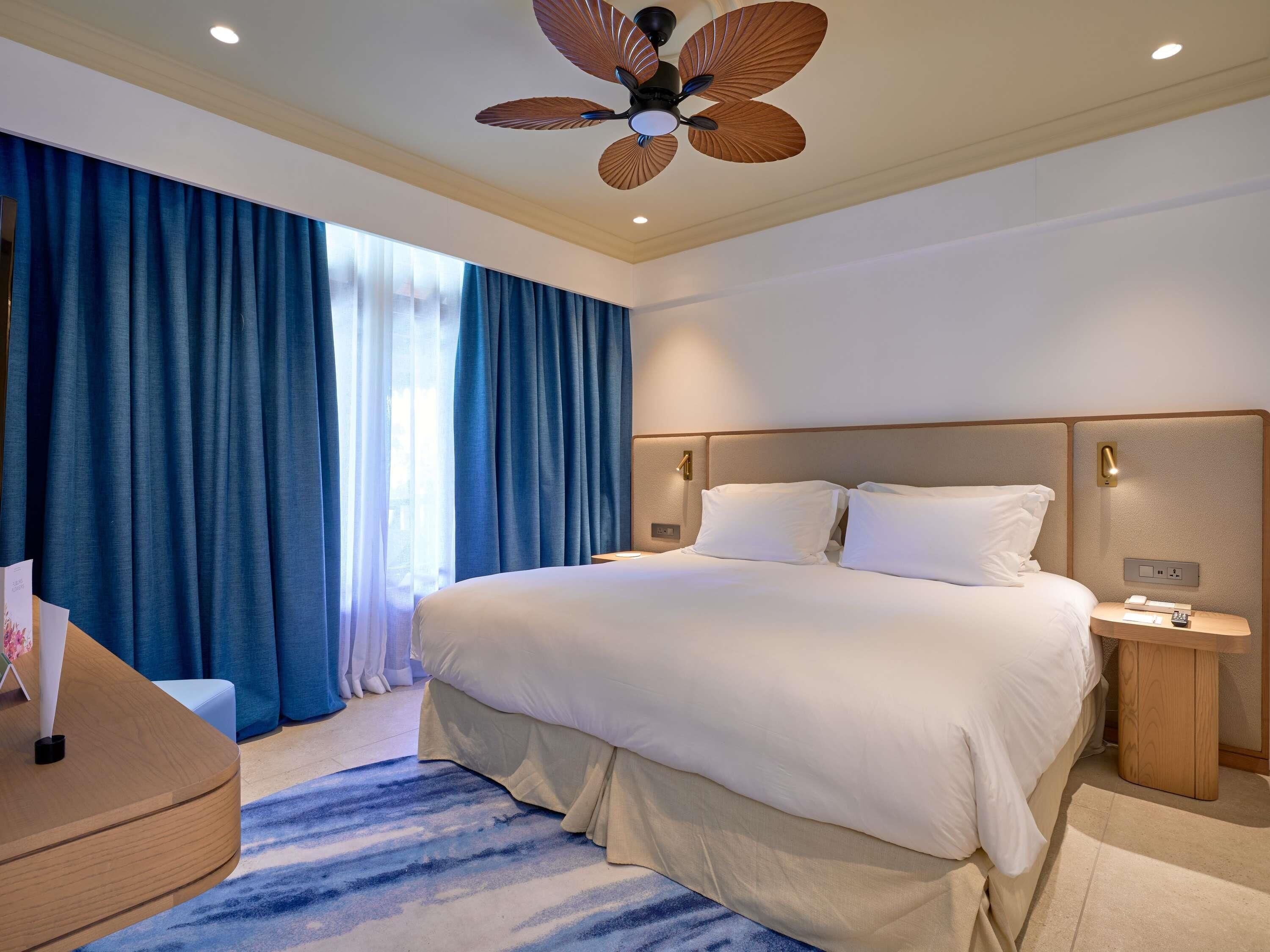 A hotel room with a large bed, a wooden nightstand, a desk, and a ceiling fan.