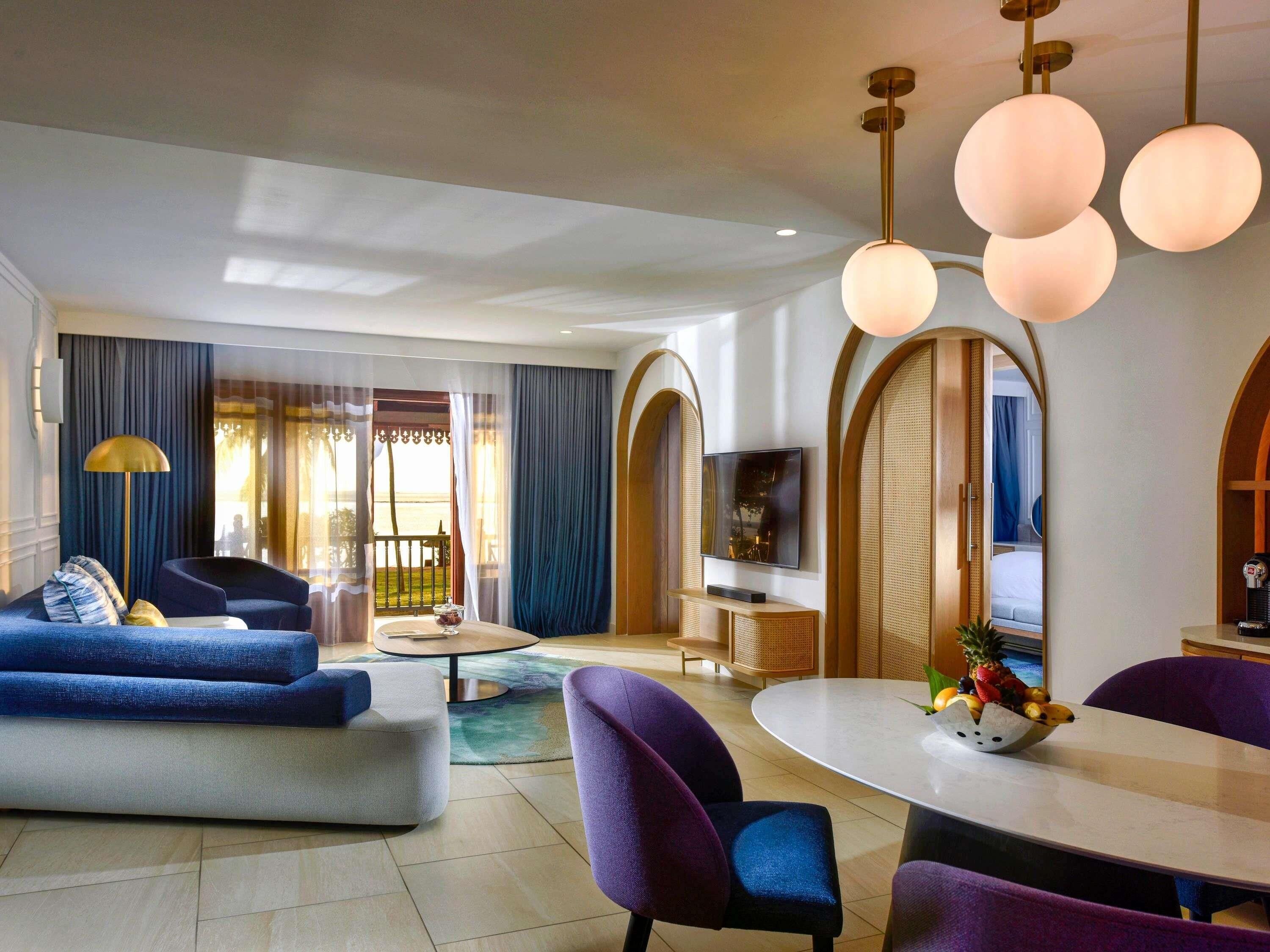 A modern hotel room with a sofa, dining table, TV, and a view of the outdoors.