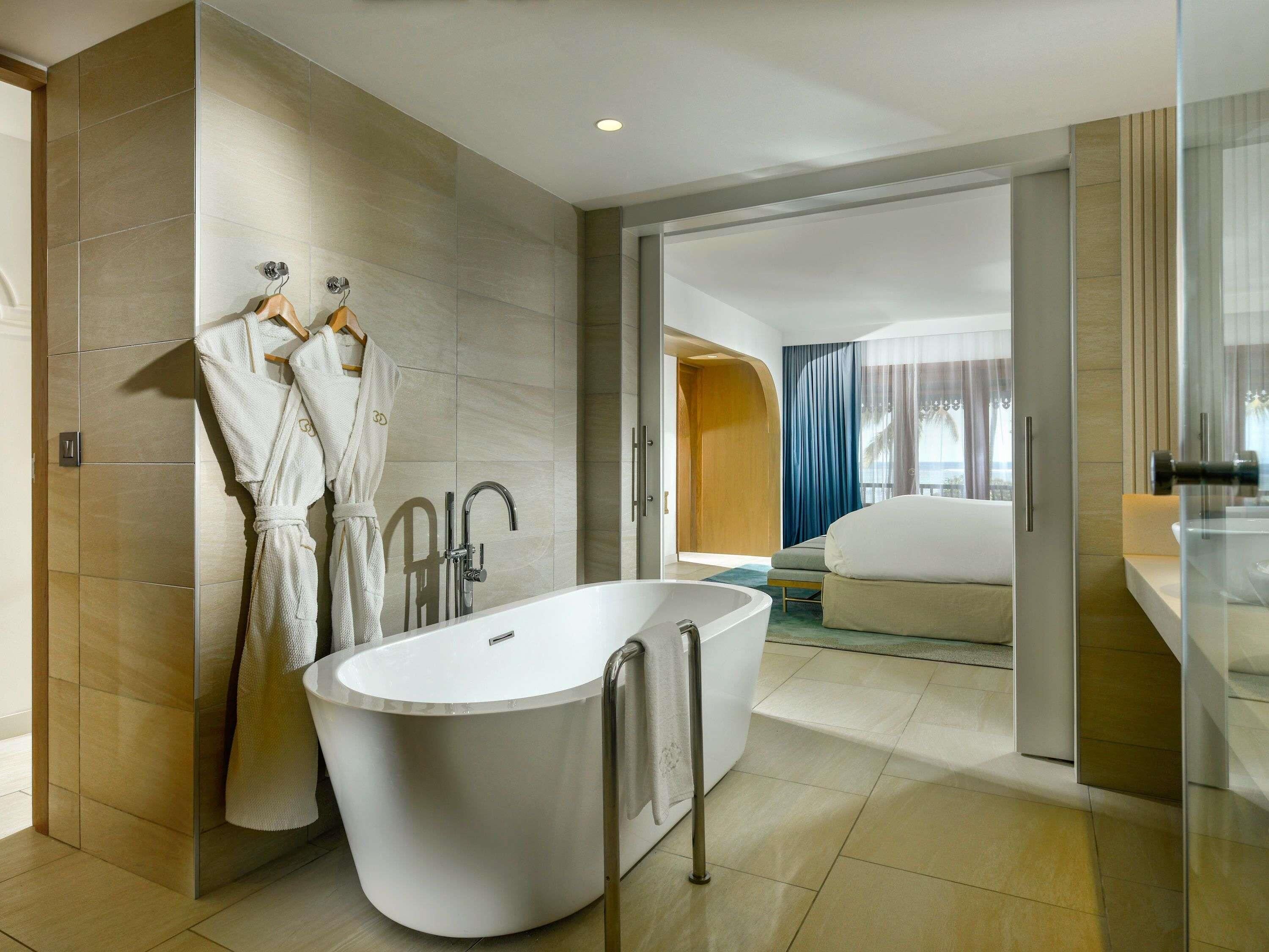 Een moderne badkamer met een vrijstaand bad, een handdoekrek met badjassen, en een zichtbare deur naar een slaapkamer.