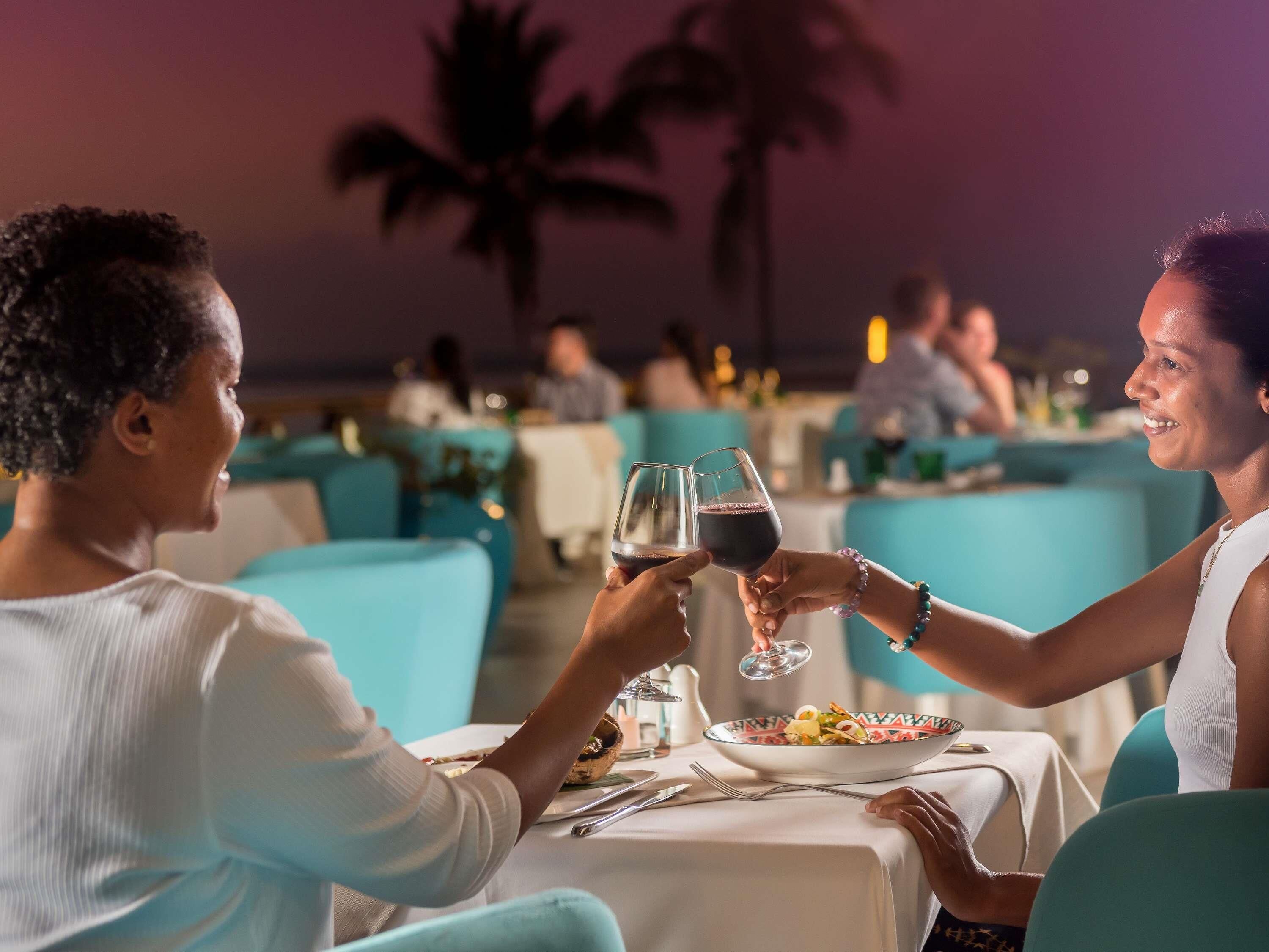 Una pareja brindando con copas de vino en una mesa con vistas a palmeras y otros comensales al fondo.