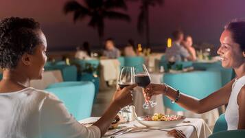 Una pareja brindando con copas de vino en una mesa con vistas a palmeras y otros comensales al fondo.