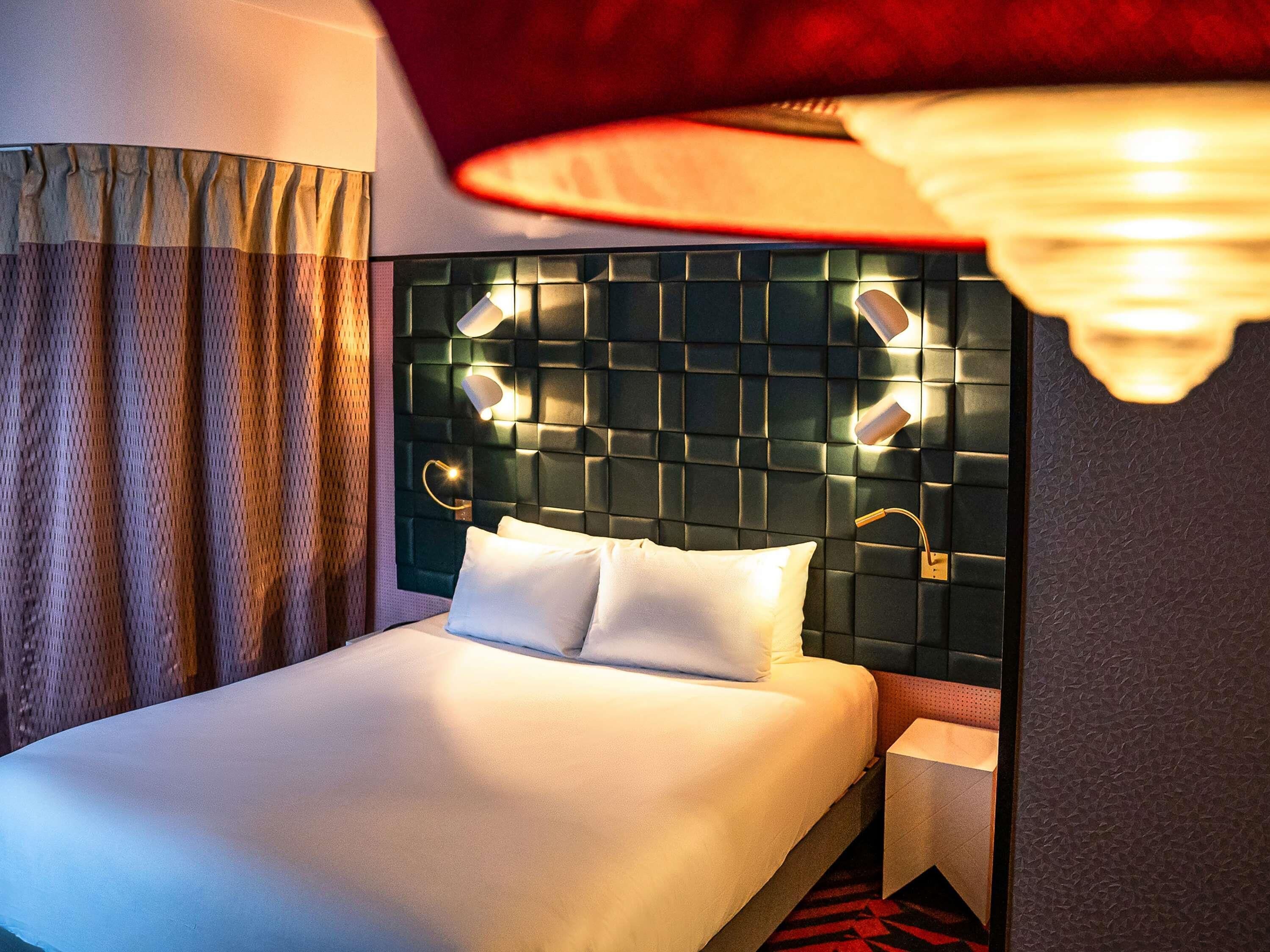 Une chambre d’hôtel avec un lit bien fait, une tête de lit dotée d’un éclairage intégré et un rideau rouge.
