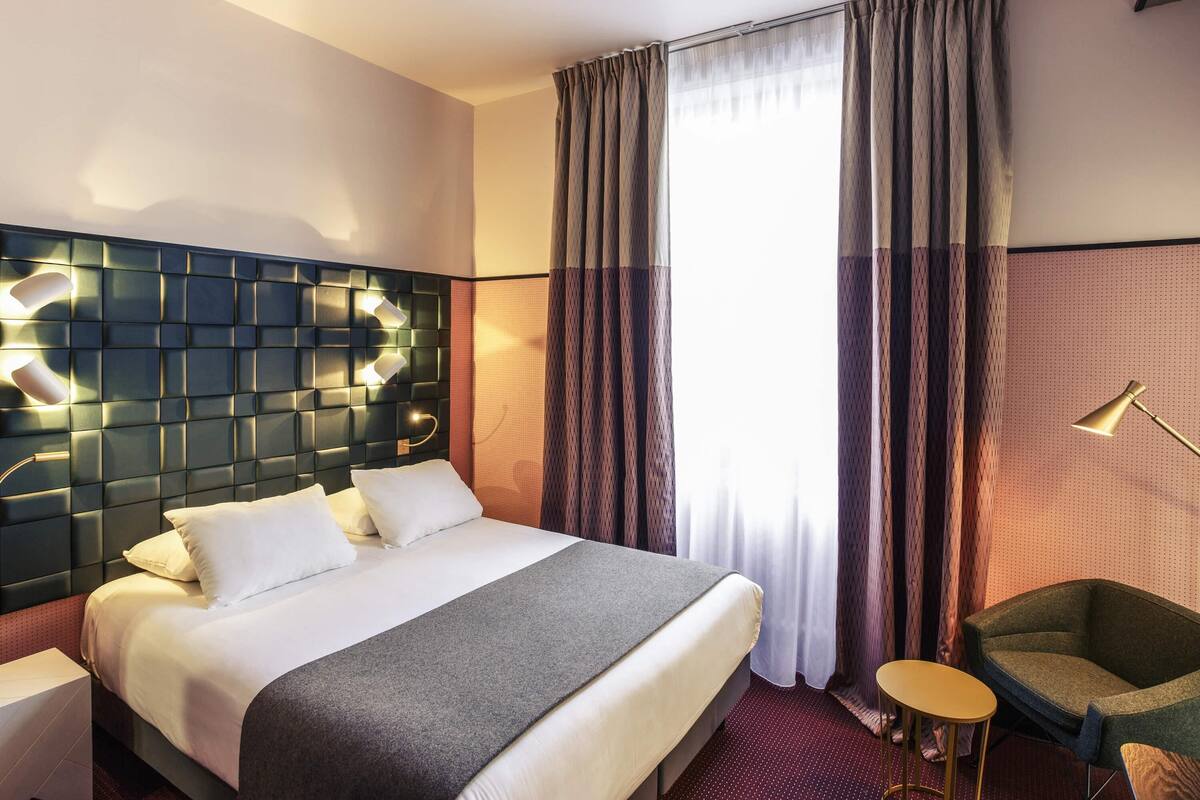 Una habitación de hotel con cama, cabecera con iluminación integrada, una silla, una mesita y una ventana con cortinas.