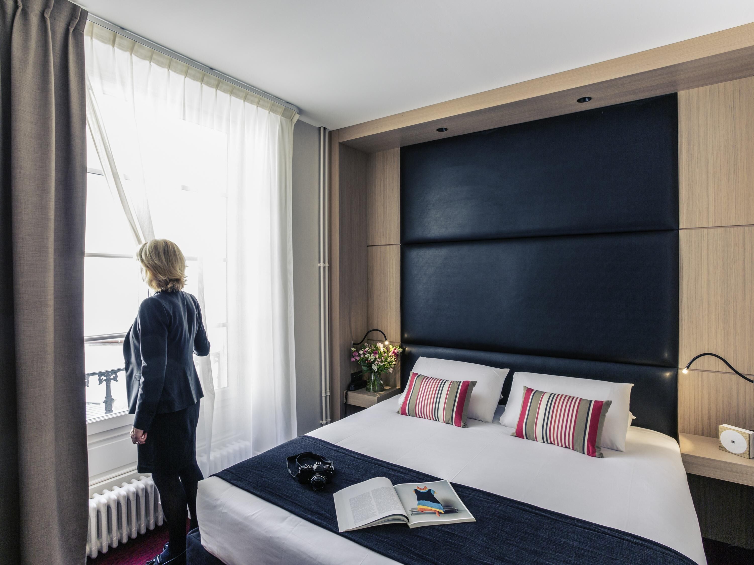 A hotel room with a large bed, a nightstand, a window with curtains, and a view of the outside.