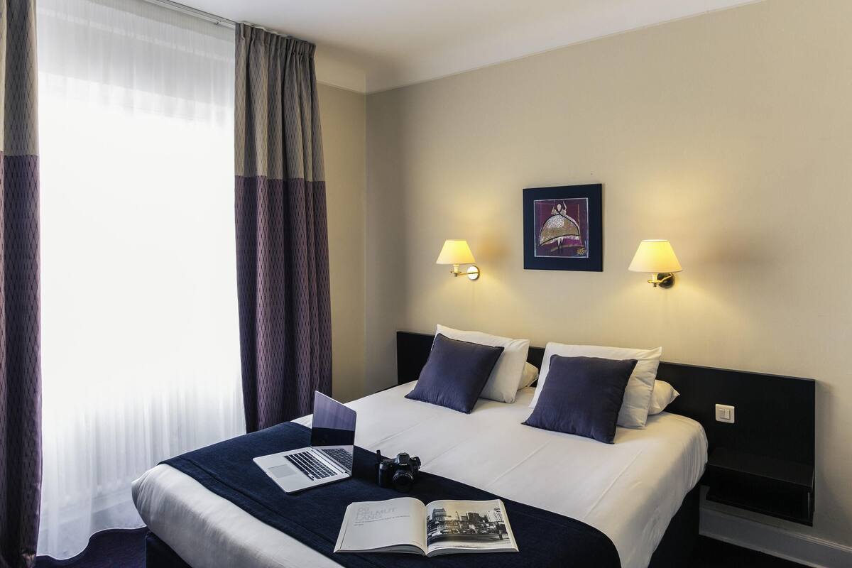 Una habitación de hotel con una cama, dos lámparas de noche, un cuadro en la pared y un ordenador portátil sobre la cama.