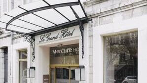 Exterior do Le Grand Hotel, com tapete vermelho na entrada, porta de vidro e varanda de metal.