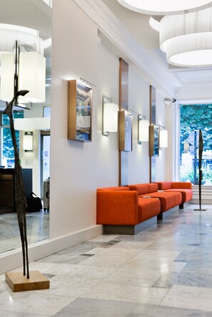 A hotel lobby with a reception desk, a row of orange seating, and a large window with a view of greenery. - Mercure Bayonne Centre Le Grand Hotel (Bayonne)