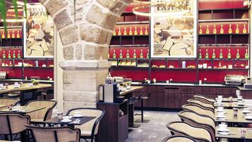 A restaurant with wicker chairs, tables, and a stone pillar. There are shelves with bottles and a poster on the wall.