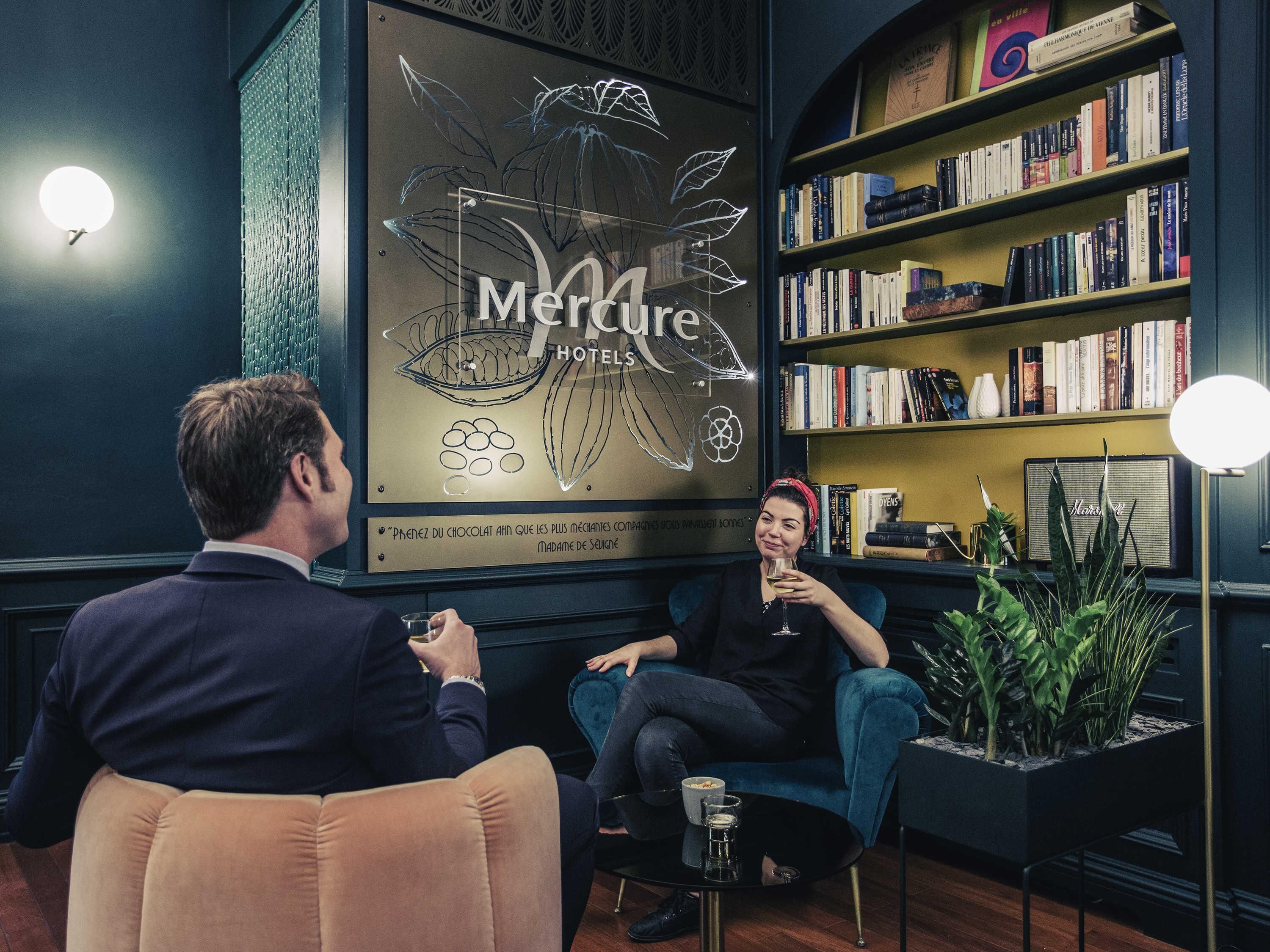 Un lobby de hotel acogedor con un cartel grande de 'Mercure Hotels', una estantería llena de libros y una planta en maceta.