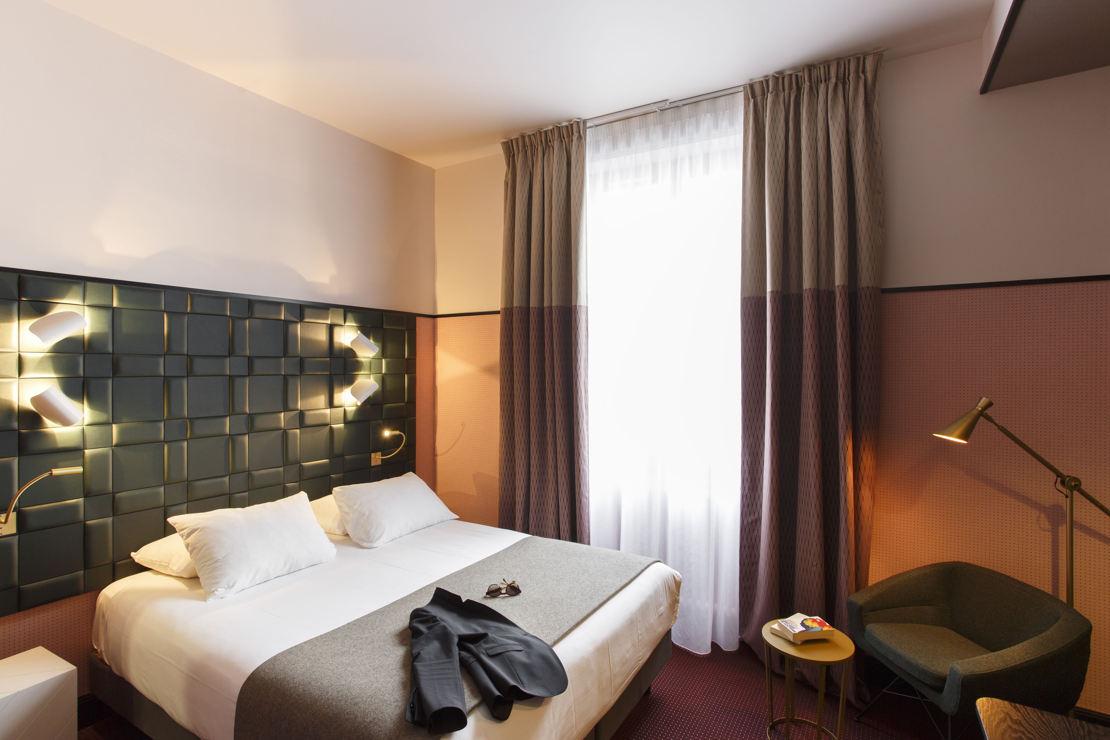 A hotel room with a large bed, a headboard with integrated lighting, a small table with a lamp, a chair, and a window with curtains.