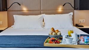 Cama bem arrumada com lençóis brancos, bandeja com café da manhã incluindo croissants, frutas e café, e mesa de cabeceira com telefone e luminária.