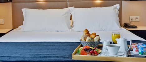 Una cama bien hecha con sábanas blancas, una bandeja con desayuno que incluye cruasanes, fruta y café, y una mesita de noche con un teléfono y una lámpara.